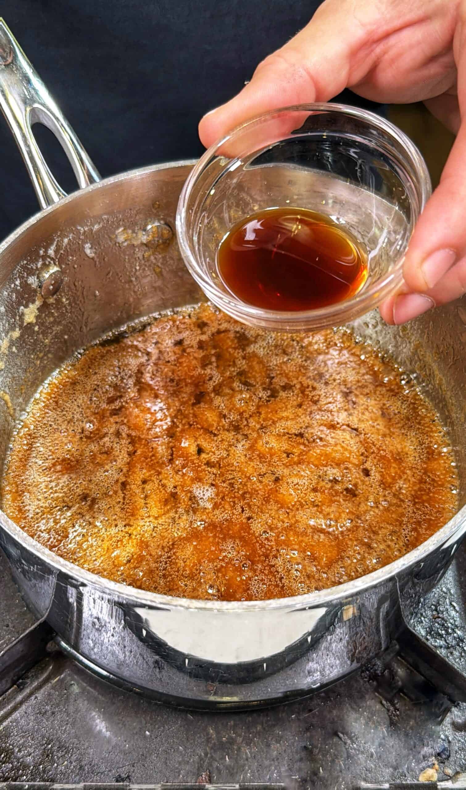 A hand holds a small glass bowl with dark liquid above a pot of bubbling brown sauce or syrup, cooking on a stovetop.