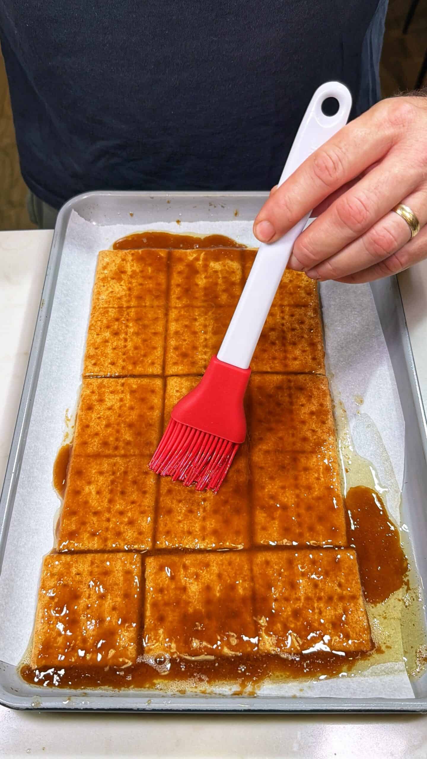 A hand uses a red silicone brush to spread sauce over rectangular pieces of tofu arranged on a parchment-lined baking sheet.