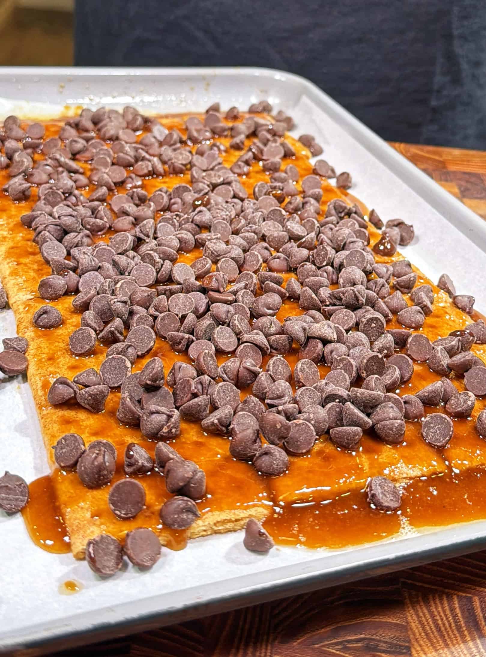 A sheet of baked pastry on parchment paper is topped with melted caramel and a generous layer of chocolate chips on a baking tray.