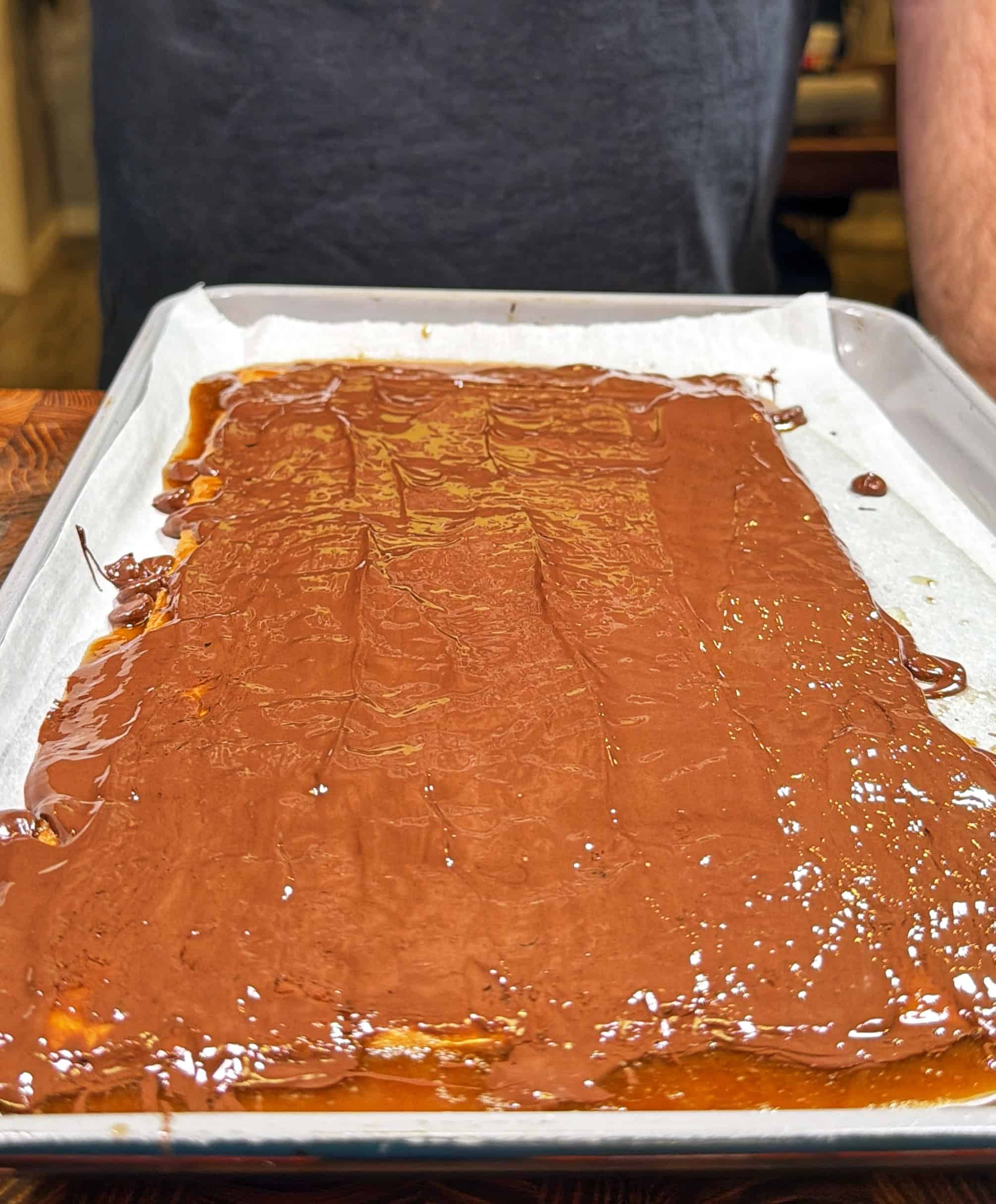 A baking tray lined with parchment paper is covered with a thin, glossy layer of melted chocolate. A person in a dark shirt stands behind the tray.