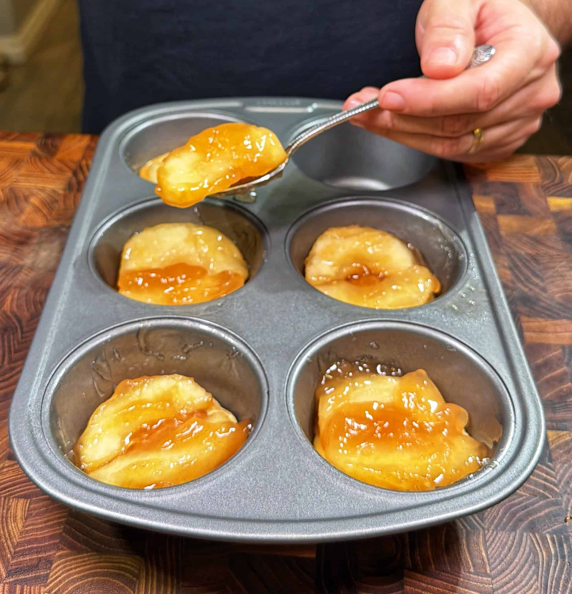 A hand uses a spoon to add glossy pie filling into a muffin tin lined with dough, preparing mini pies on a wooden countertop.