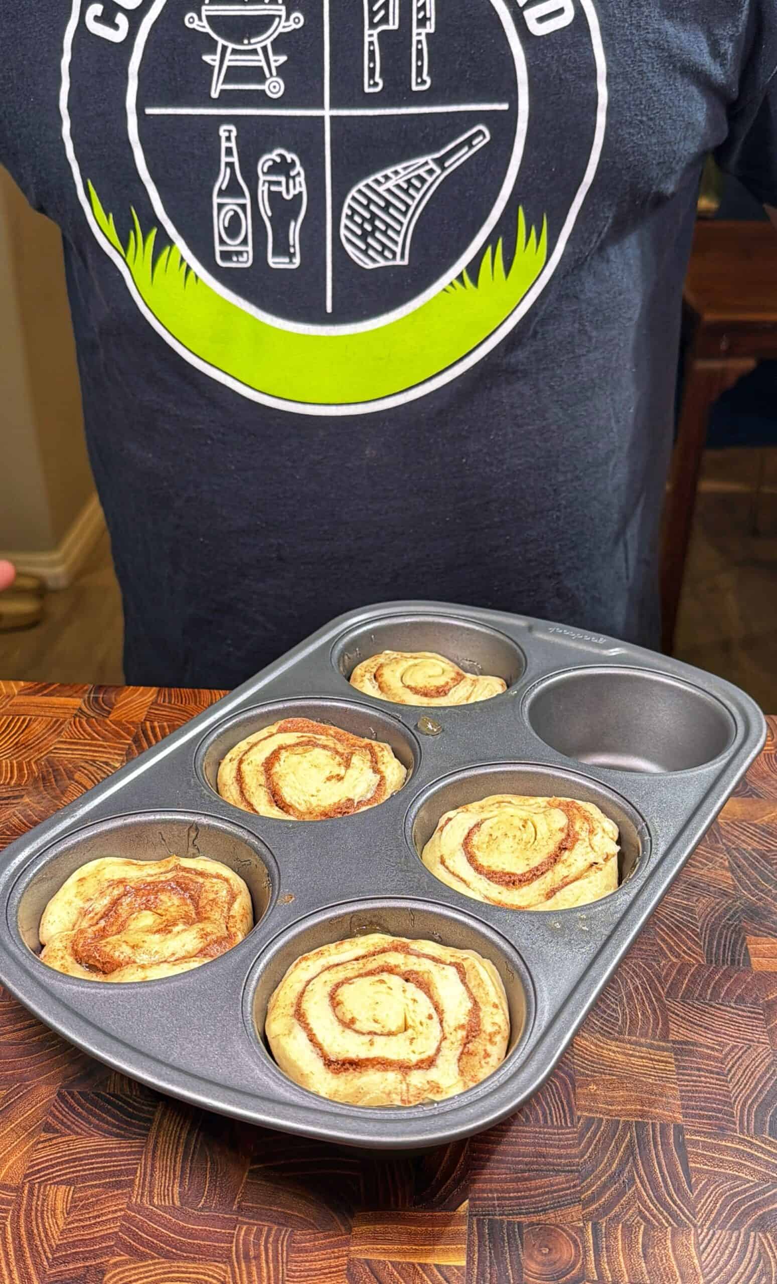 A person wearing a black shirt with a grilling graphic holds a muffin tin containing four large cinnamon rolls on a wooden countertop.