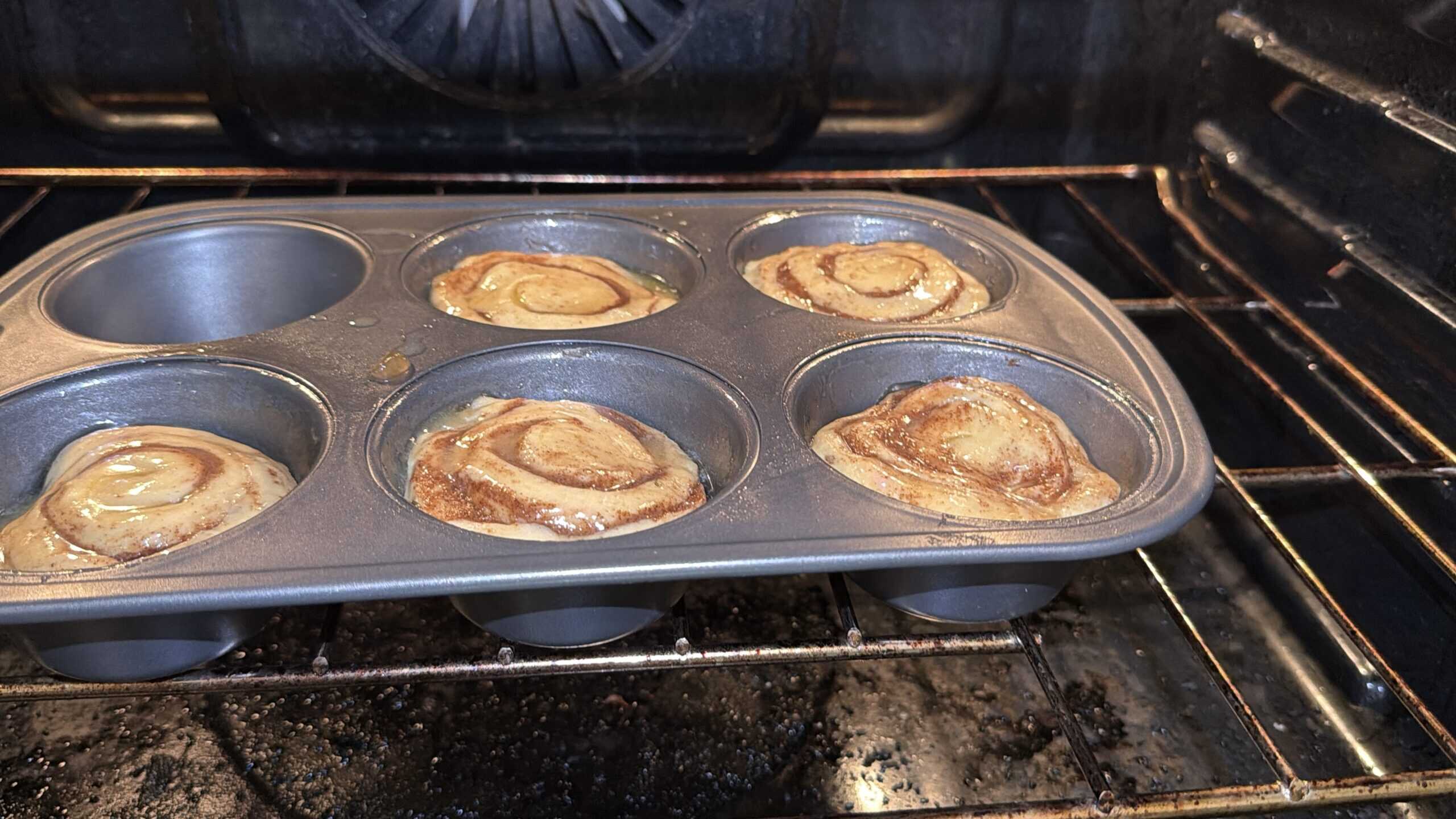 A metal muffin tin with six spaces, four filled with cinnamon rolls baking in an oven. The cinnamon rolls are golden brown and rising, with swirled tops visible.