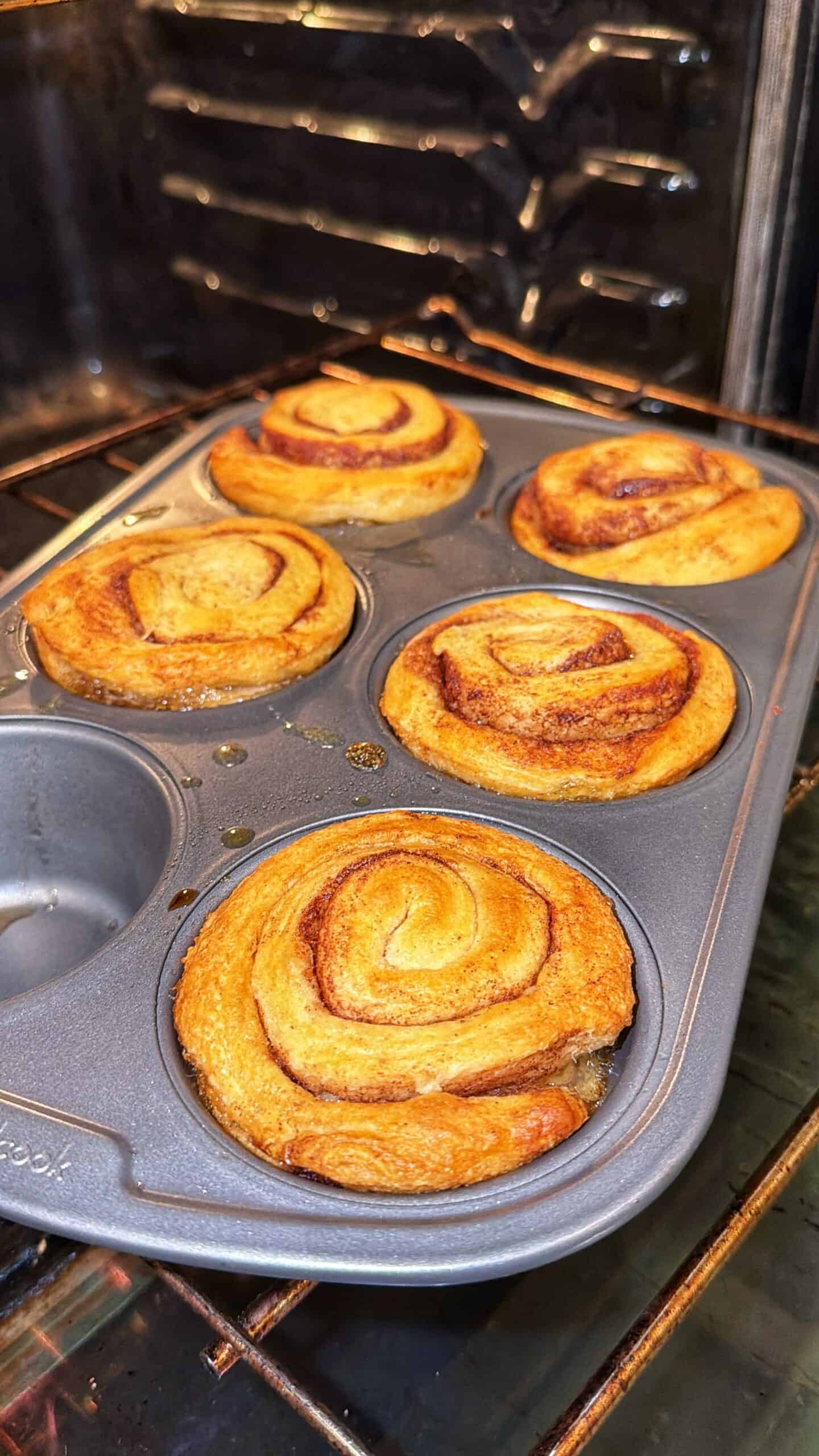 Six golden-brown cinnamon rolls baking in a muffin tin inside an oven. The rolls are puffed and slightly crisp on top, with swirls of cinnamon visible. One tin compartment is empty.