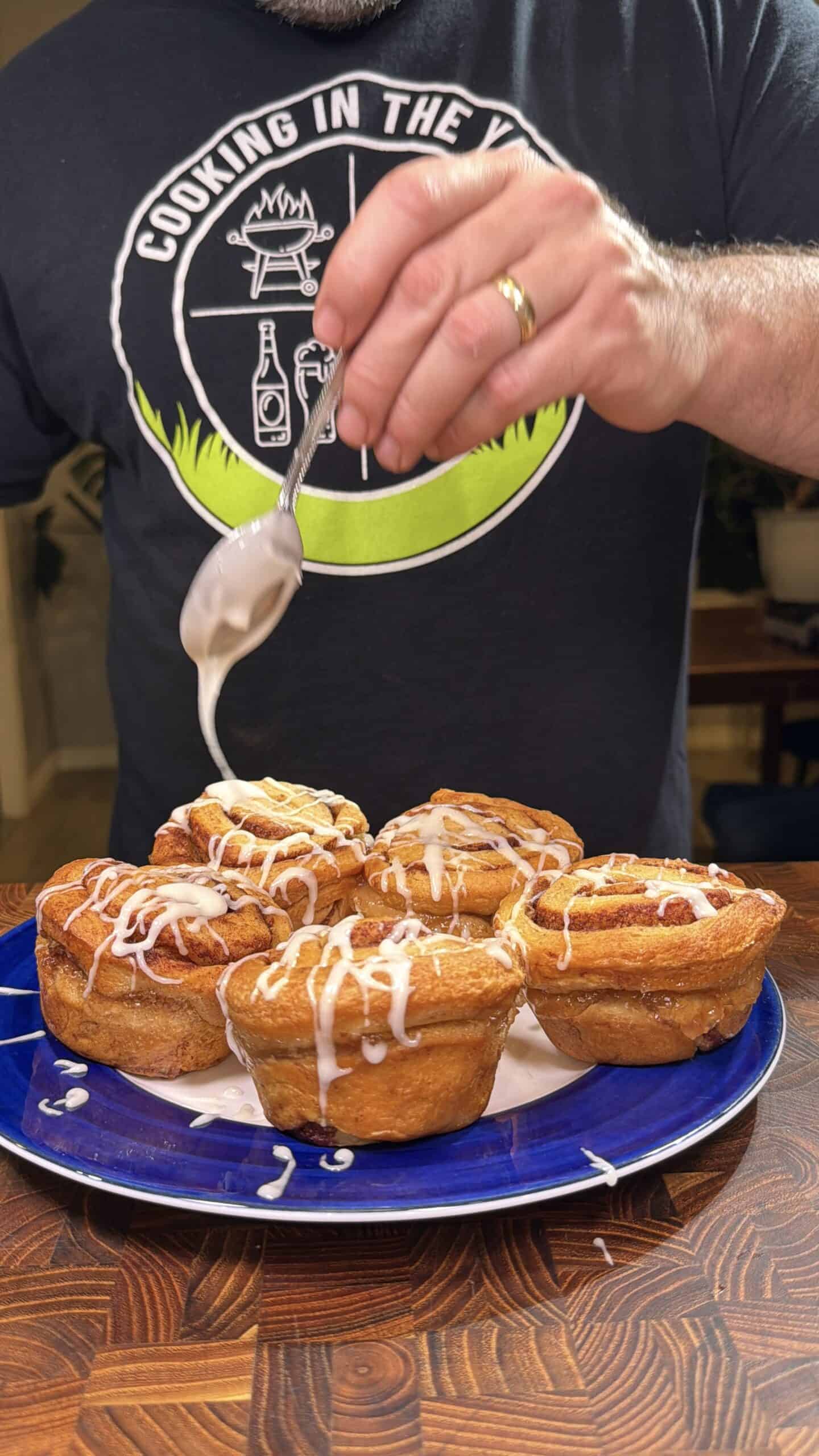 A person in a Cooking in the Yard shirt drizzles icing with a spoon over six cinnamon rolls on a blue plate, set on a wooden table.