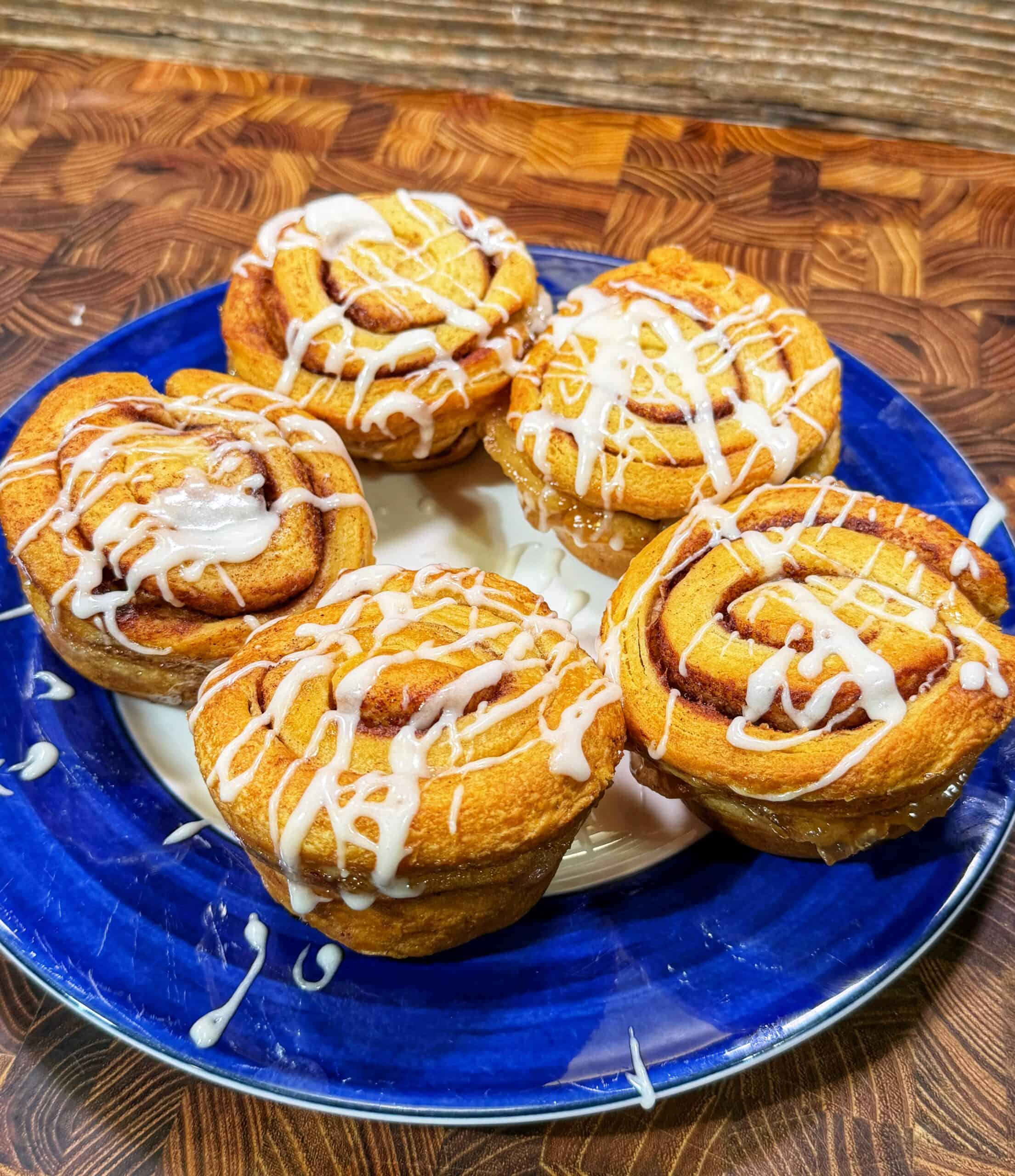 Four cinnamon rolls topped with white icing are arranged on a blue plate, which sits on a wooden surface. The rolls appear freshly baked and golden brown.