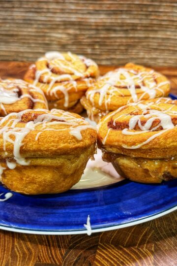 Four cinnamon rolls with white icing drizzled on top sit on a blue plate, placed on a wooden surface with a textured brown background.