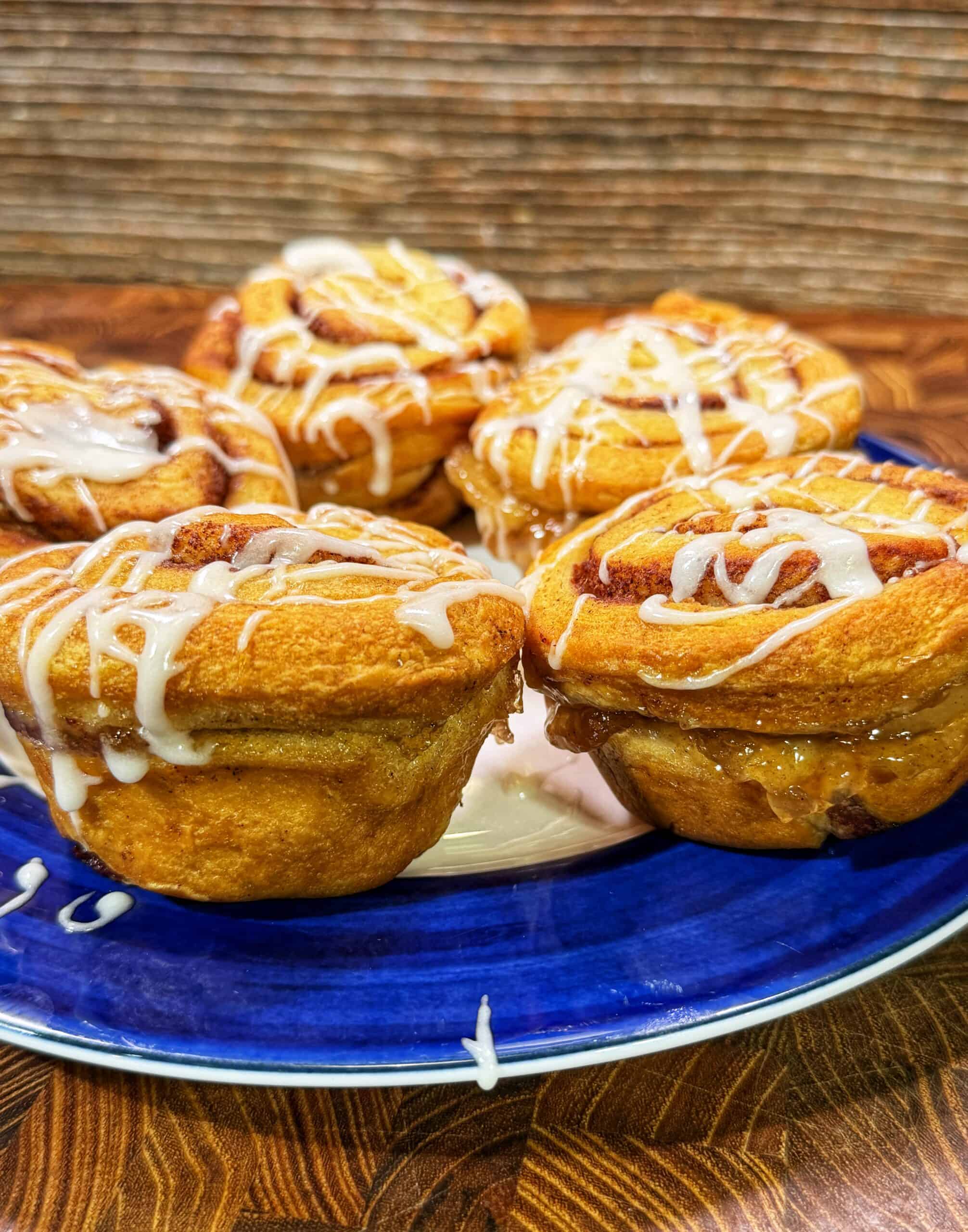 Four cinnamon rolls with white icing drizzled on top sit on a blue plate, placed on a wooden surface with a textured brown background.