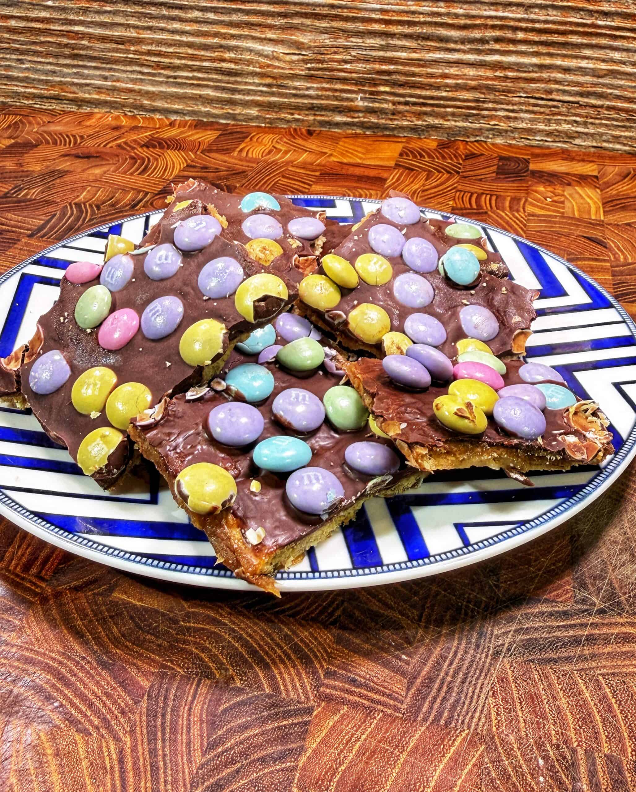A plate with four pieces of chocolate bark topped with pastel-colored candy-coated chocolates, placed on a patterned wooden surface.