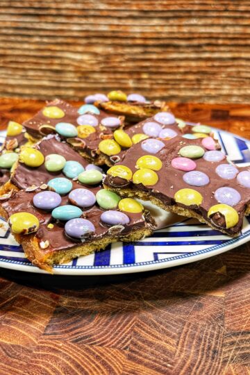 A plate holds pieces of chocolate bark topped with colorful pastel candy-coated chocolates, set on a wooden surface with a textured brown background.