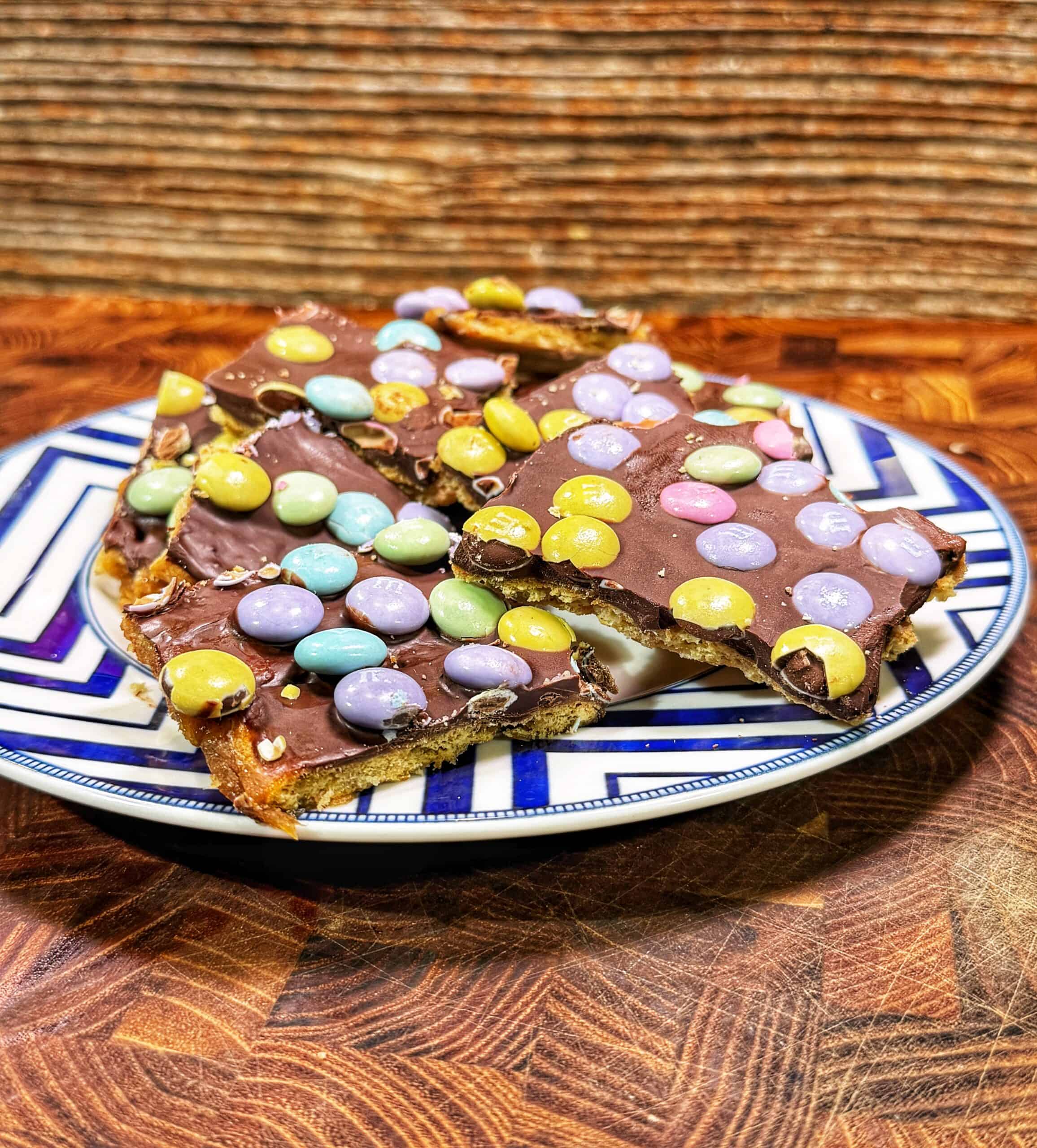 A plate holds pieces of easter crack topped with colorful pastel candy-coated chocolates, set on a wooden surface with a textured brown background.