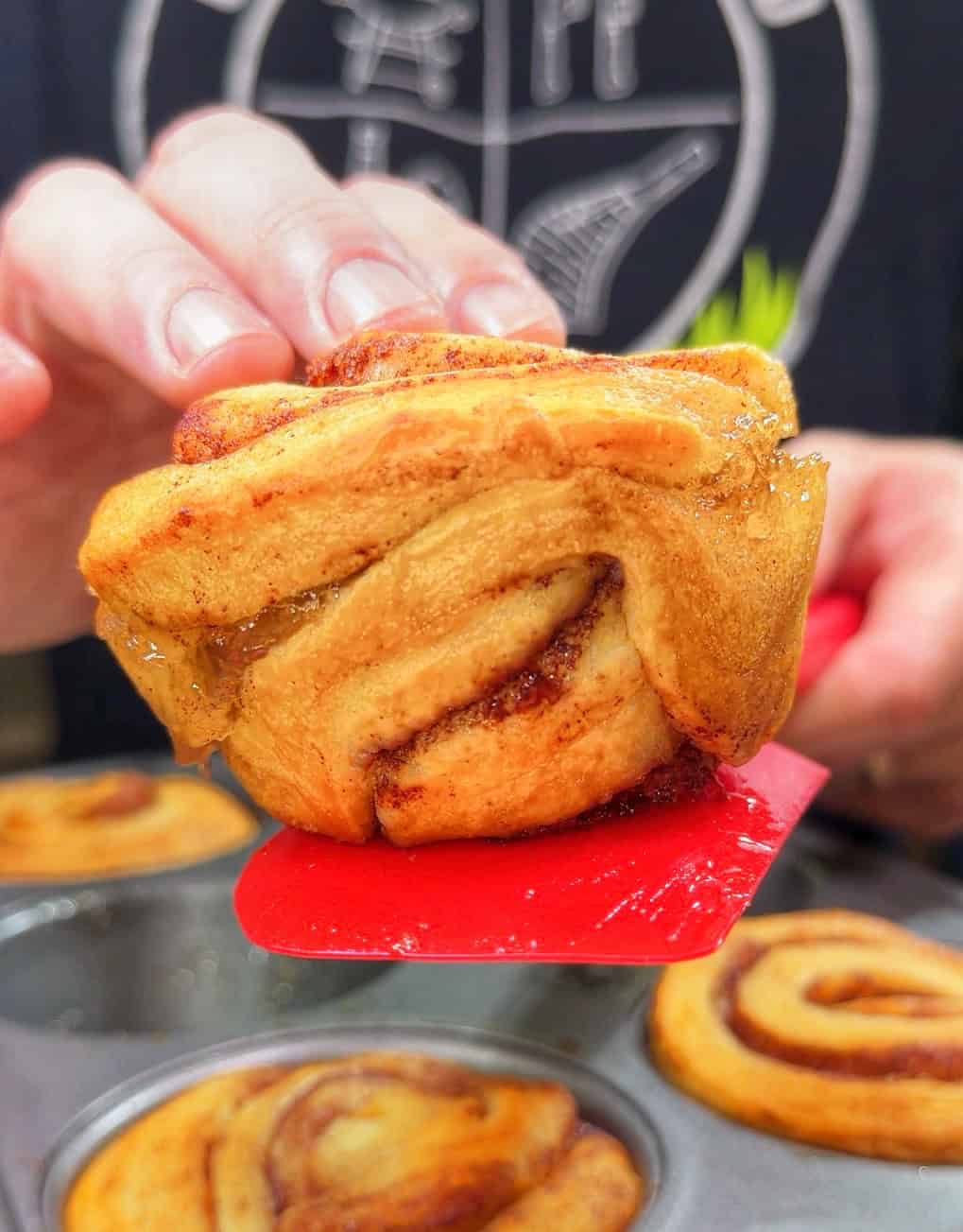 A close-up of a hand lifting a freshly baked cinnamon roll from a muffin tin with a red spatula. Other cinnamon rolls are visible in the background.
