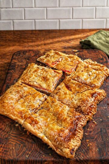 Golden, flaky pastry cut into six large squares sits on a wooden cutting board, with a tiled white background and a green cloth off to the side.