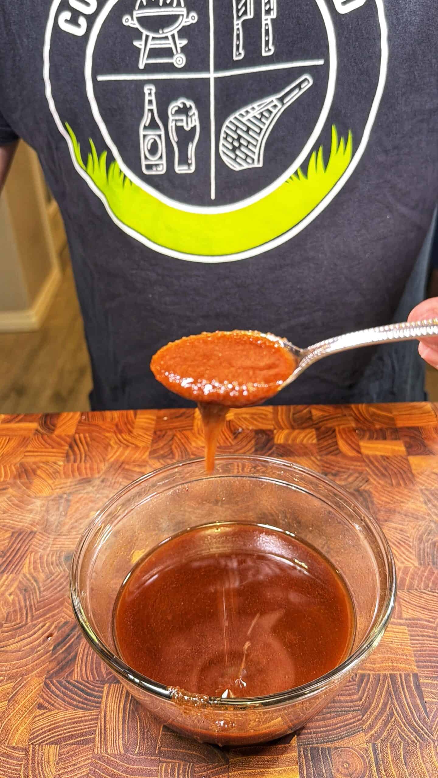 A person holds a spoonful of thick, reddish-brown barbecue sauce above a glass bowl of the same sauce, with some sauce dripping back into the bowl, on a wooden surface.