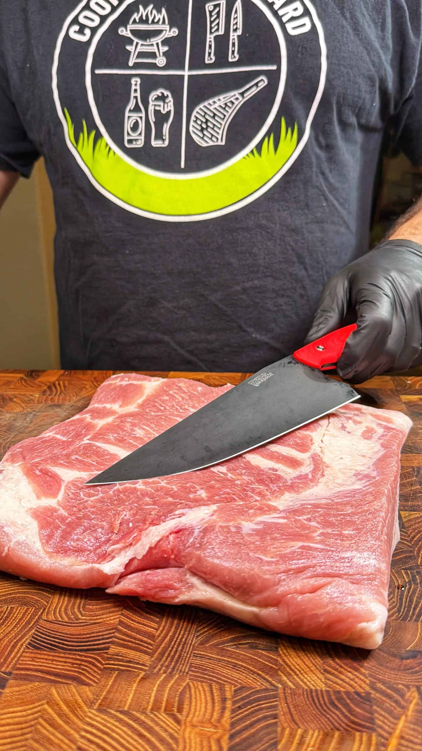 A person wearing a black glove and a “Cooking in the Yard” T-shirt slices a large piece of raw pork on a wooden cutting board with a large knife.
