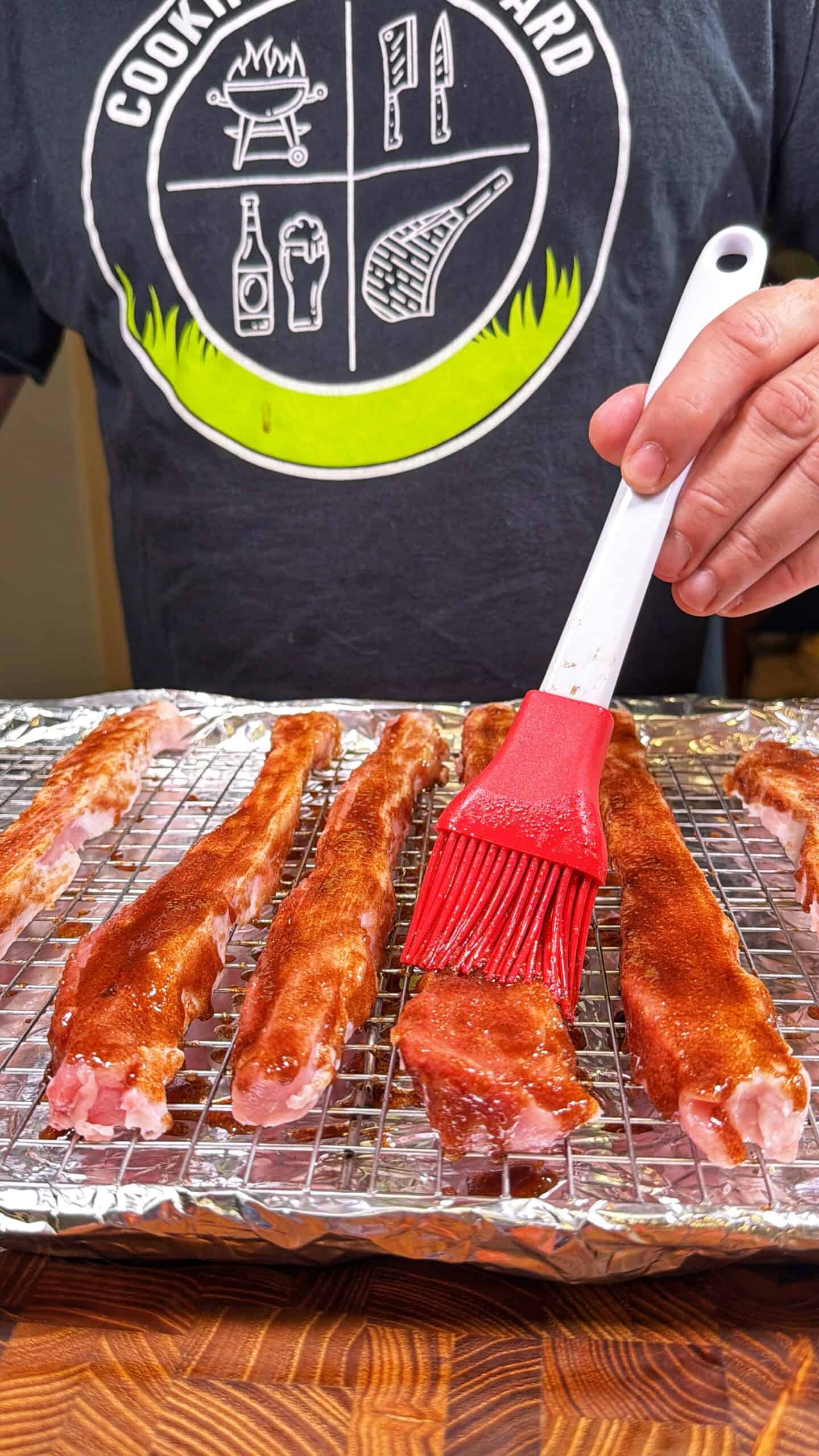 A person brushes barbecue sauce onto several raw pork ribs placed on a wire rack over a foil-lined baking sheet. The person is wearing a dark shirt with grilling-themed graphics.