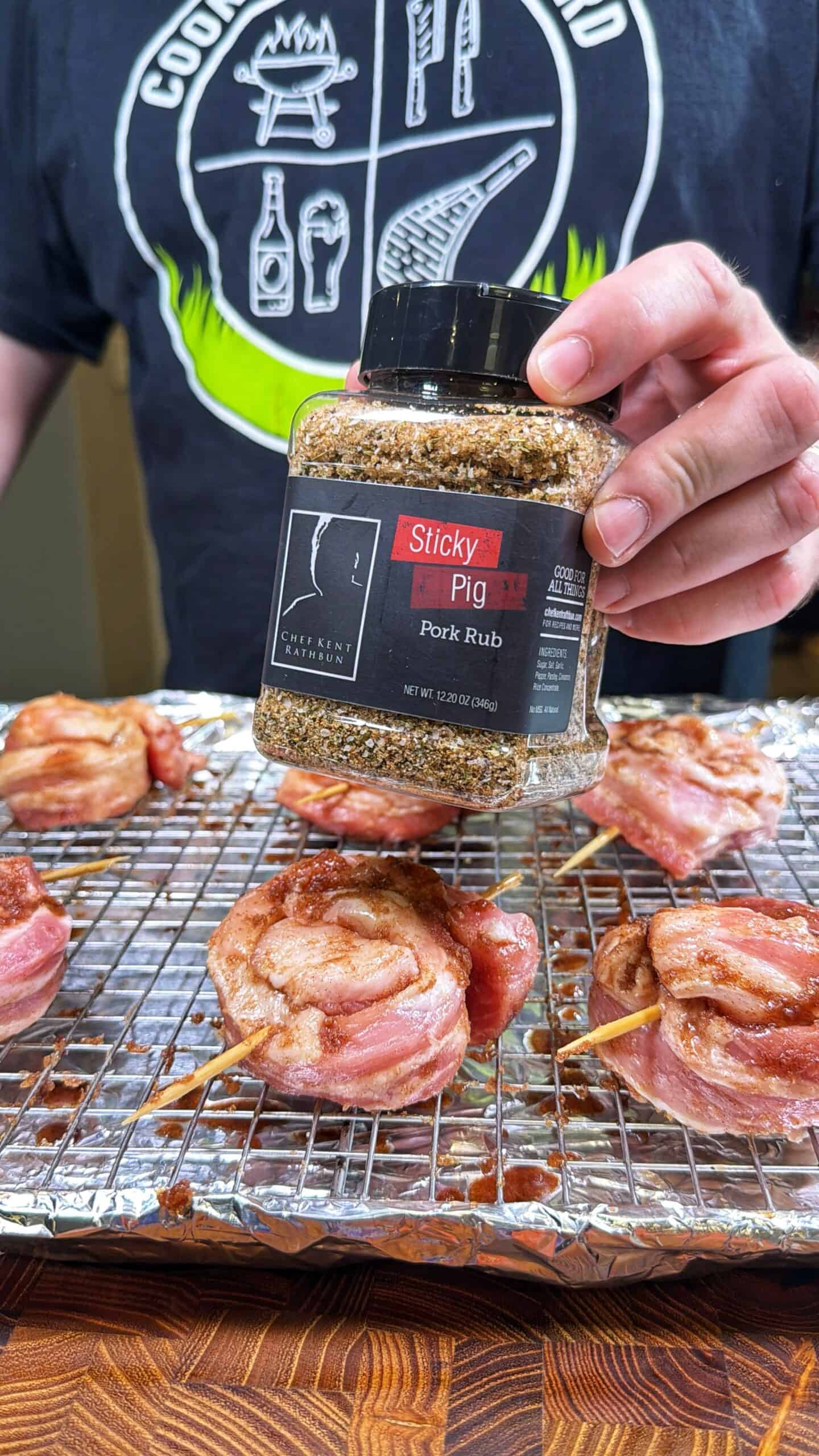 A person holds a container of Sticky Pig pork rub over bacon-wrapped meat pieces on a wire rack, ready for seasoning. The person wears a black shirt with grilling graphics.