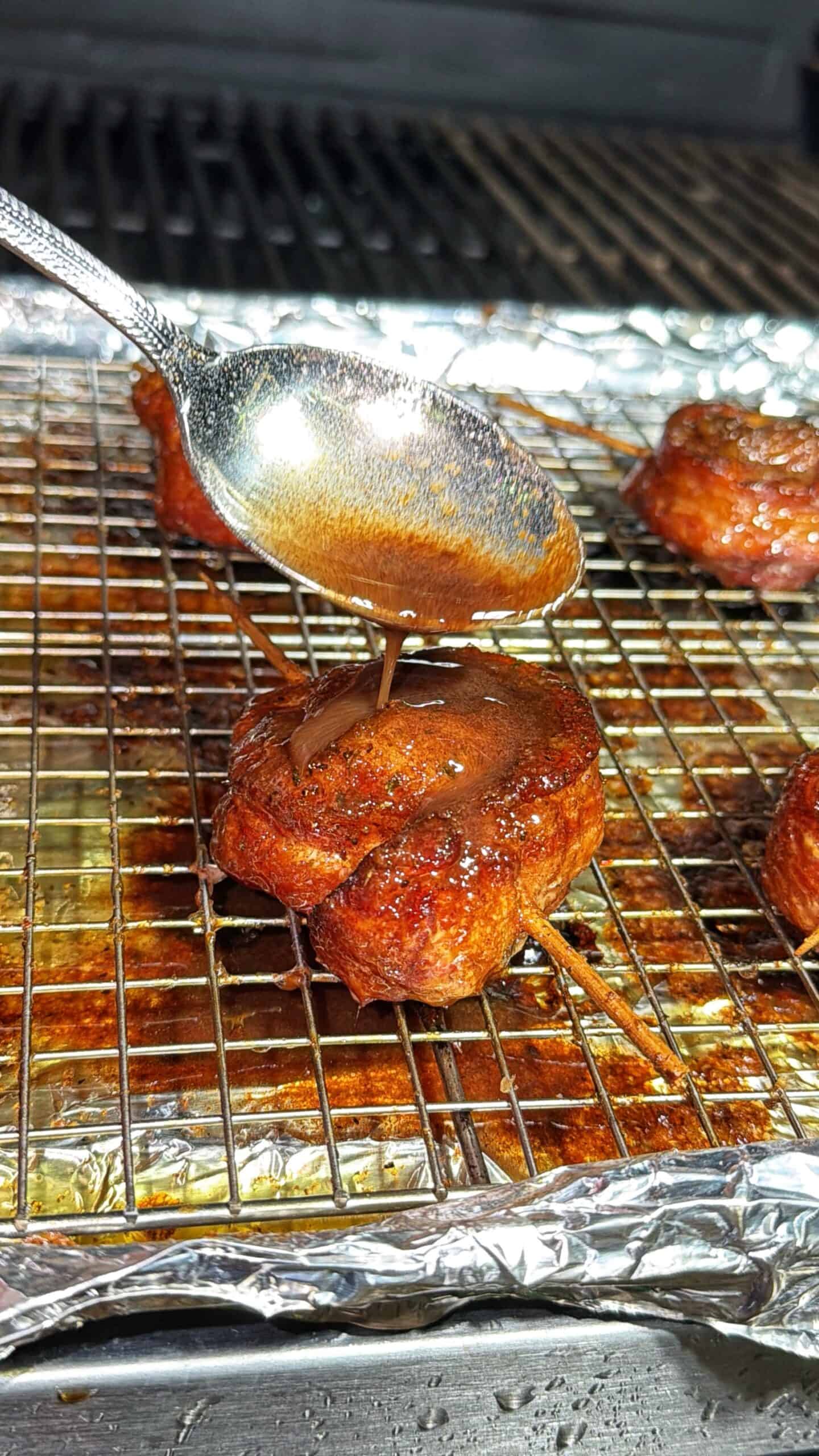 A hand holds a spoon, drizzling sauce onto a small, round, bacon-wrapped food item on a wire rack over a foil-lined baking sheet, with more pieces and a grill in the background.