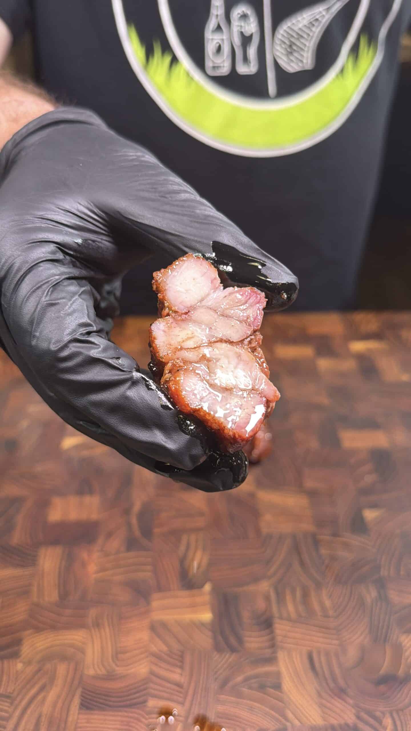 A person wearing a black glove holds up two slices of juicy, glazed, smoked meat over a wooden cutting board. A black shirt with a grilling-themed design is partially visible in the background.