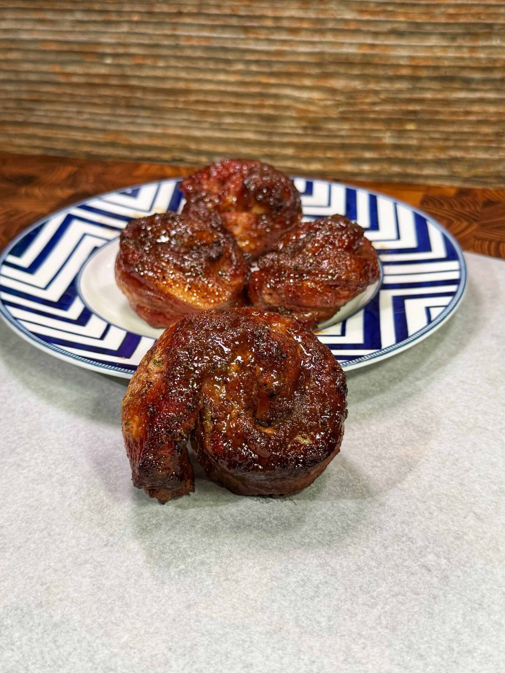 Three pieces of cooked, glazed meat shaped like cinnamon rolls are arranged on a blue and white patterned plate, with one piece set in front on white parchment paper. The background shows a rustic wooden surface.
