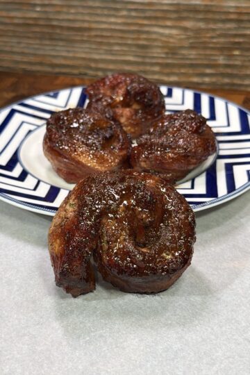 Three pieces of grilled, glazed meat shaped like cinnamon rolls are arranged on a patterned plate, with one piece in the foreground, against a wooden background.