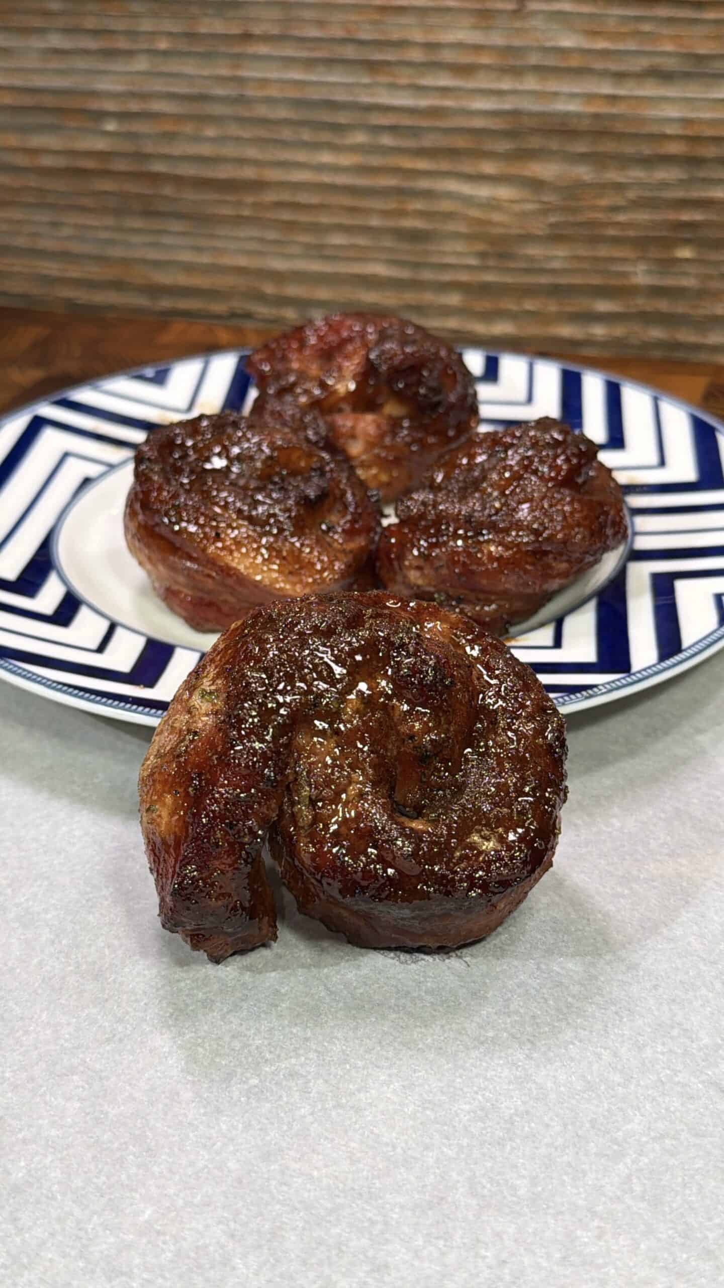 Three pieces of grilled, glazed meat shaped like cinnamon rolls are arranged on a patterned plate, with one piece in the foreground, against a wooden background.