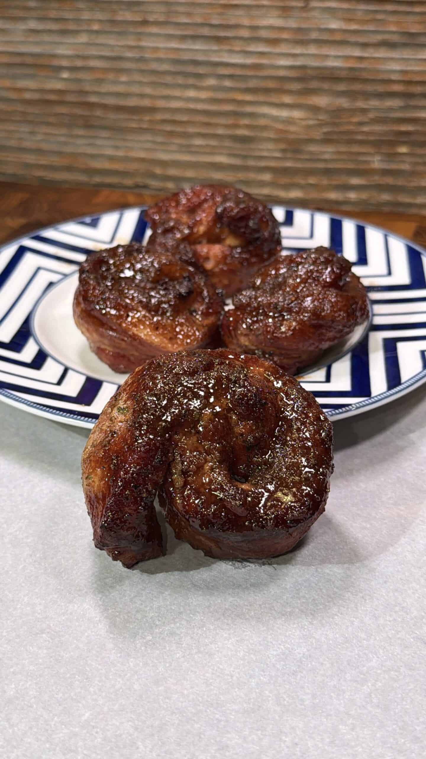 Three glazed, spiral-shaped pastries or buns are arranged on a blue and white patterned plate, with one bun placed in front on a white surface. The background is a textured, brown wall.