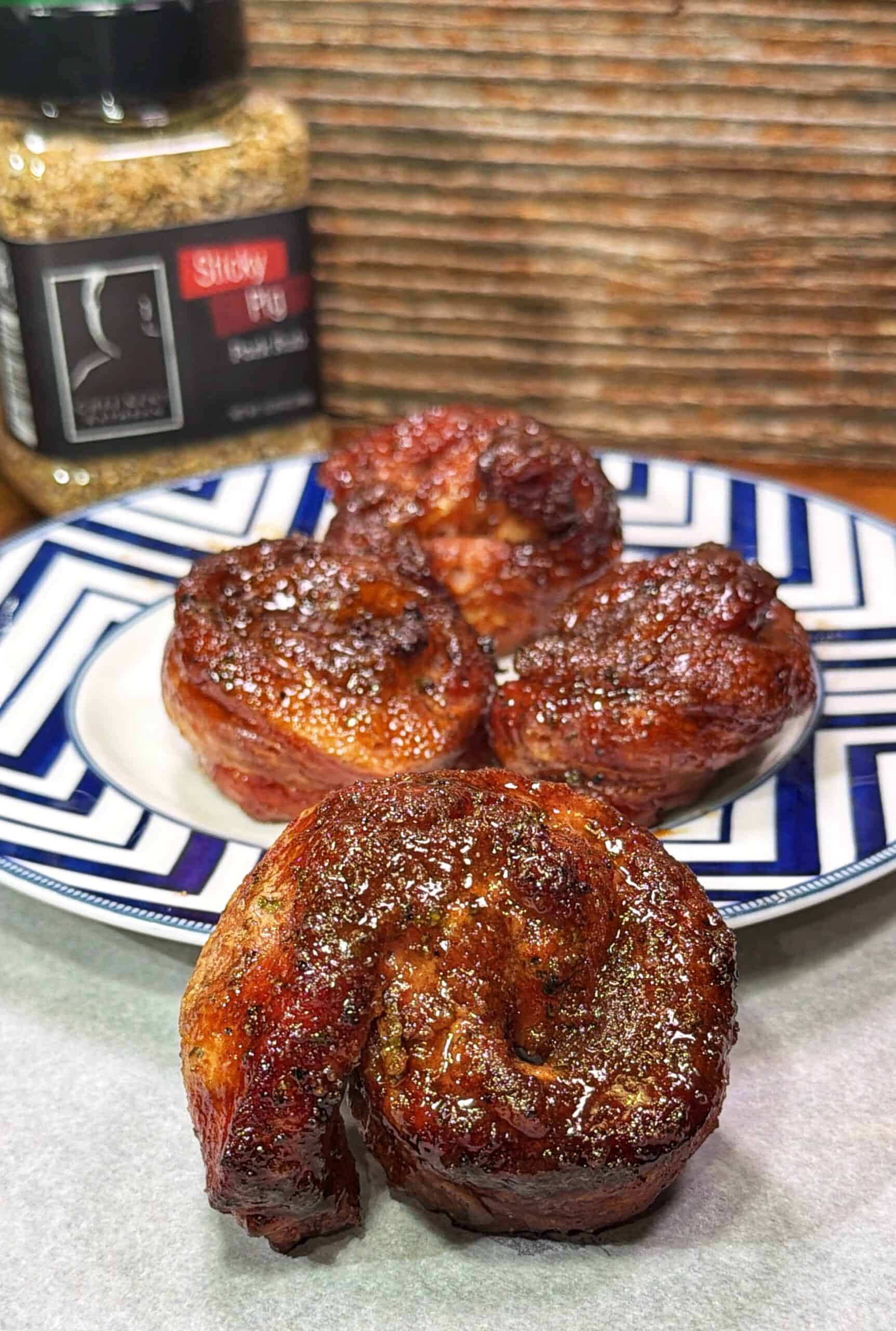 Four pieces of seasoned and cooked meat are arranged on a blue and white plate, with one piece in the foreground. A jar of Sticky Pig pork rub seasoning is in the background.