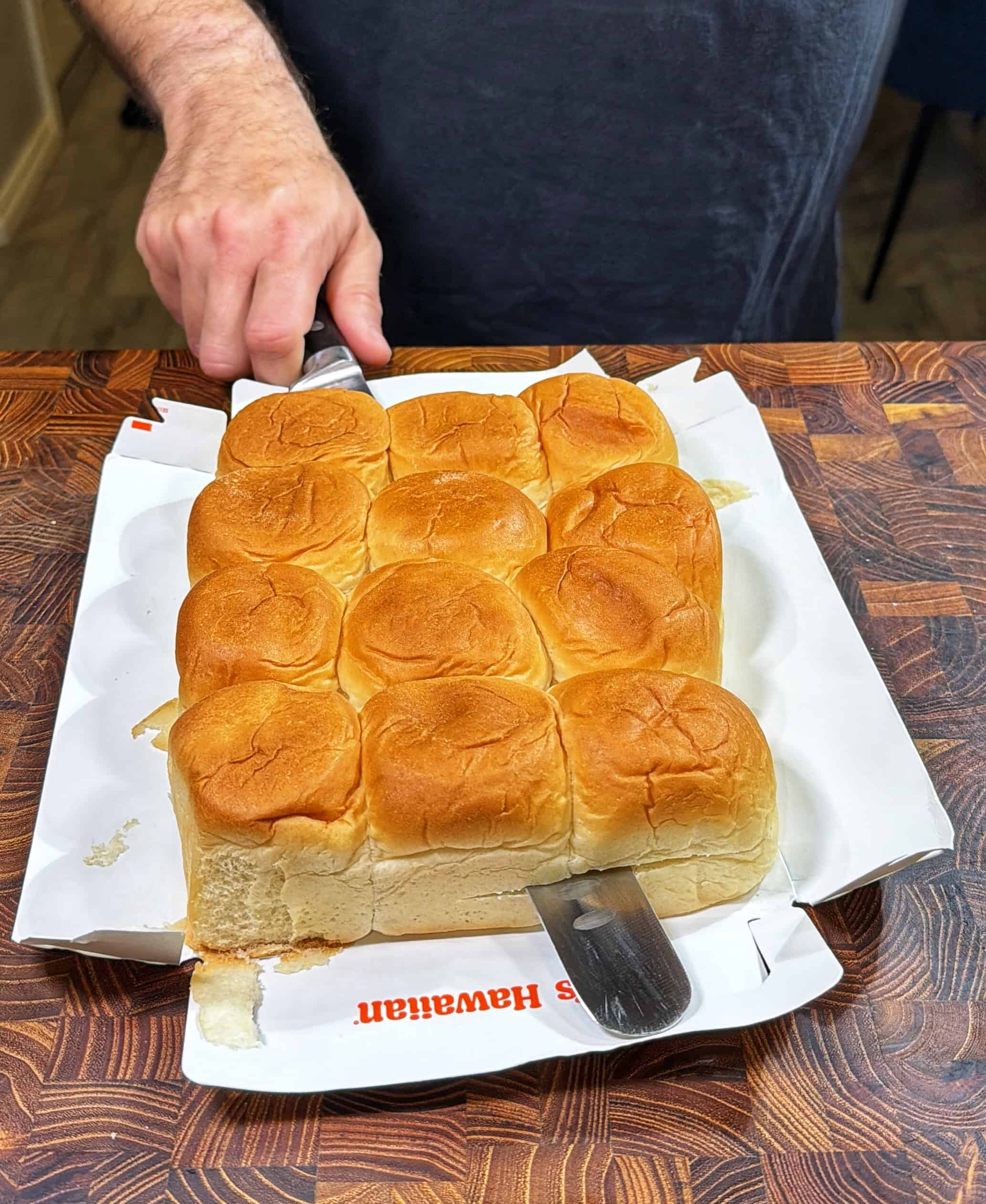 A person slices a tray of soft, golden-brown Hawaiian rolls on a wooden cutting board, using a large knife. The rolls are still attached in a rectangular block, and a paper liner is underneath.