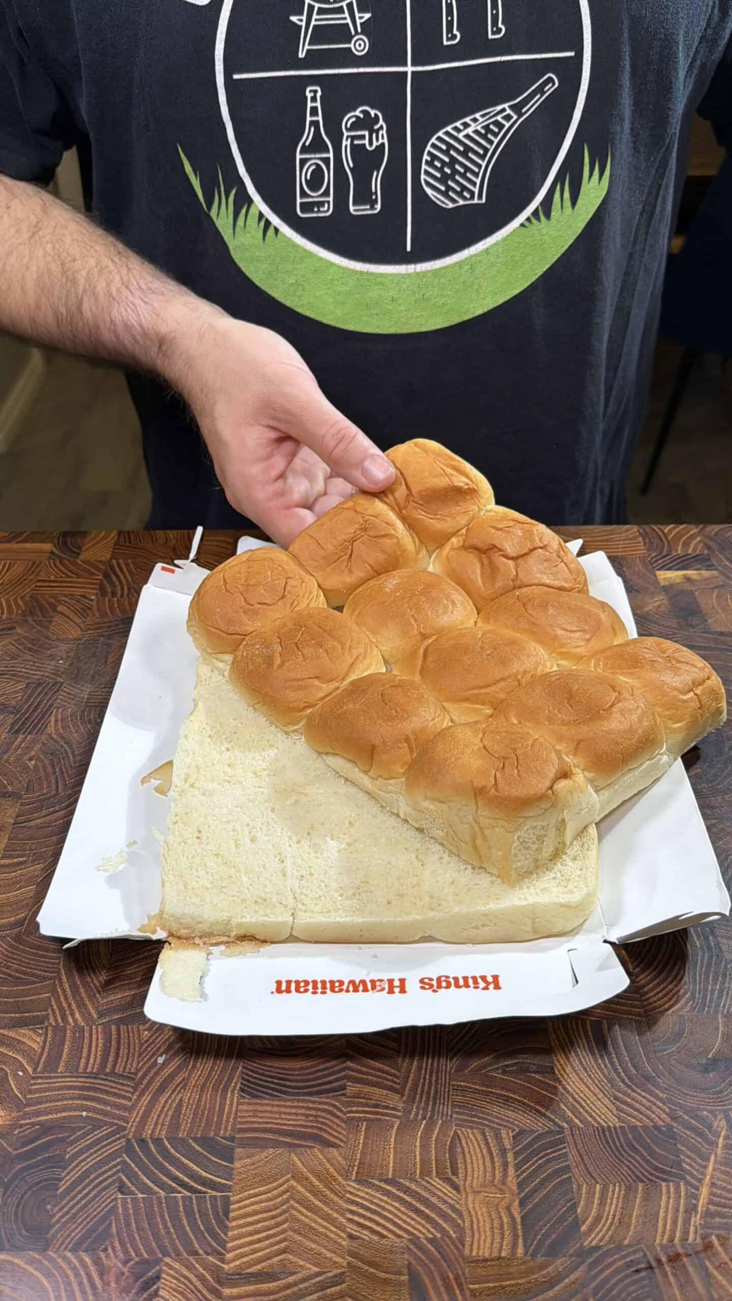 A person places a large sheet of connected hamburger buns on an opened fast food box set on a wooden table. The buns appear soft and are partially separated from the bottom half.