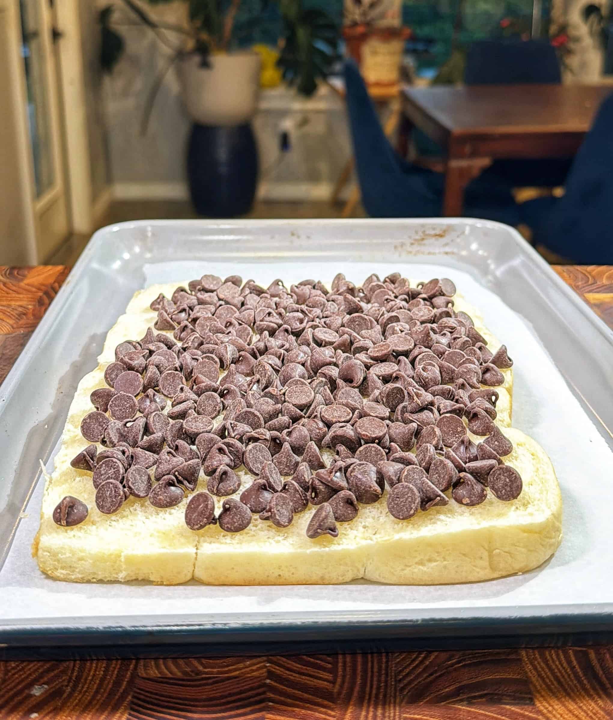 A baking tray holds a rectangular sheet of bread topped with a thick layer of chocolate chips, ready to be baked. The background shows a dining area with chairs, a table, and some plants.