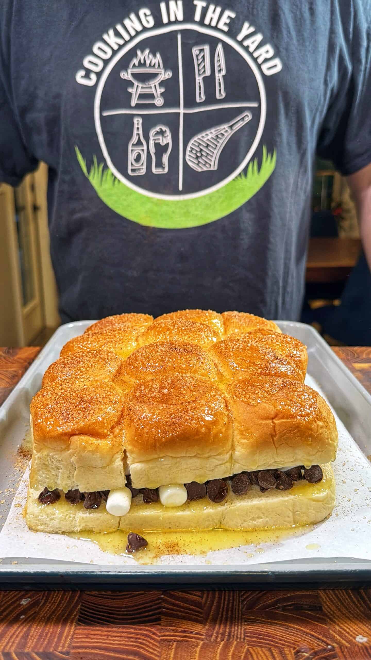 A close-up of a tray with pull-apart rolls filled with chocolate chips and marshmallows, in front of a person wearing a Cooking in the Yard t-shirt with barbecue-themed graphics.