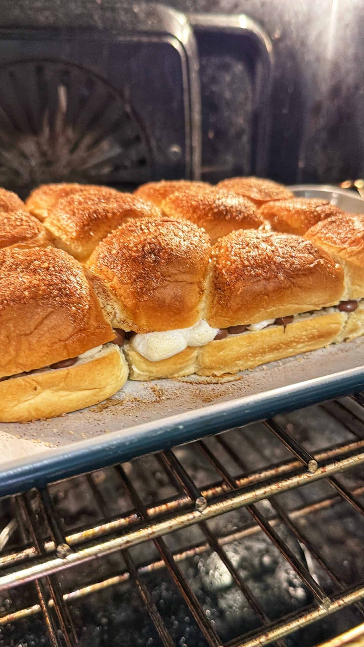 A tray of golden-brown slider sandwiches baking in the oven, with melted cheese or marshmallow oozing from the sides of the soft buns on a metal baking sheet.