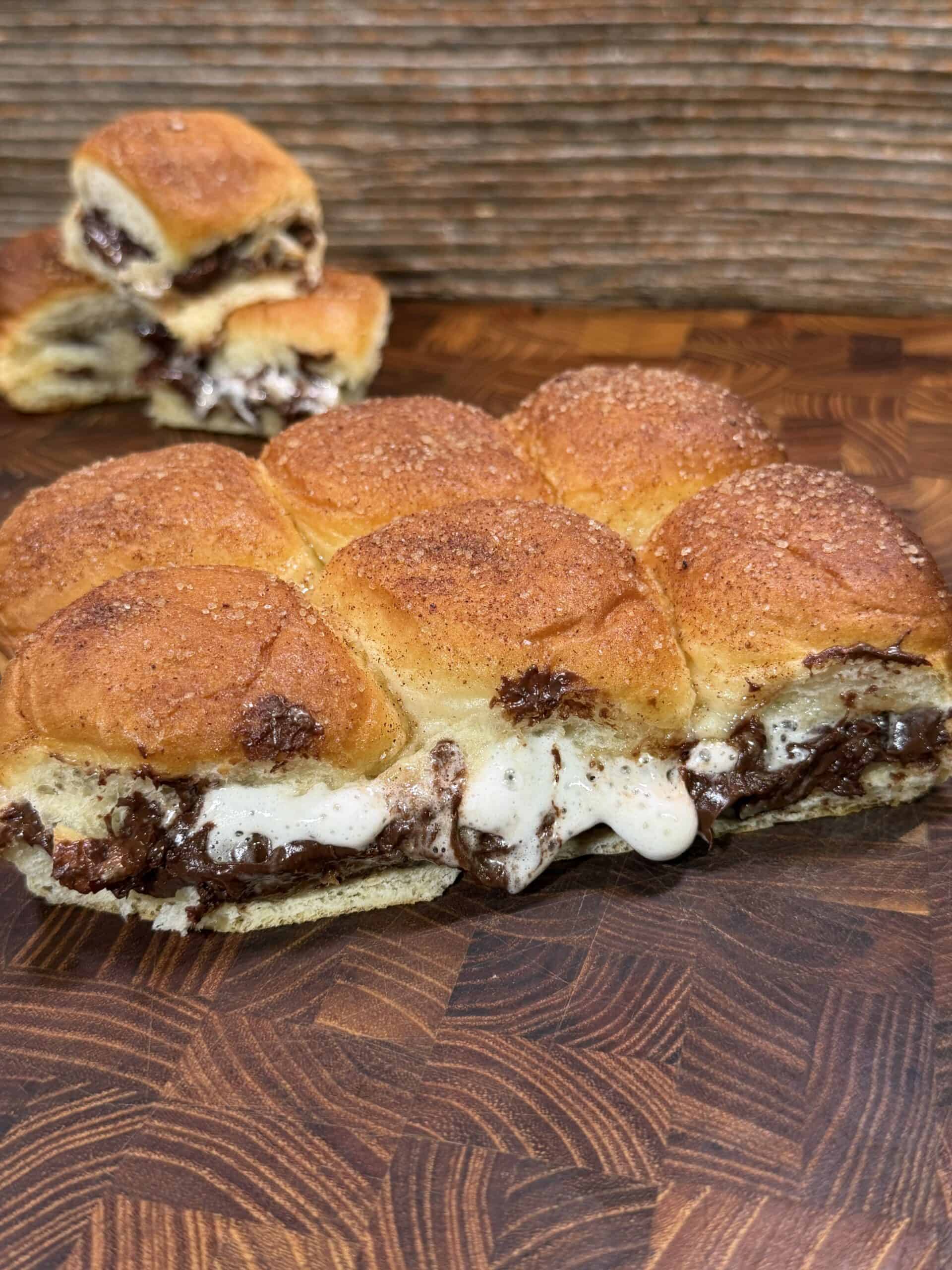 A close-up of sweet pull-apart rolls filled with melted chocolate and gooey marshmallow on a wooden surface, with some buns separated in the background.