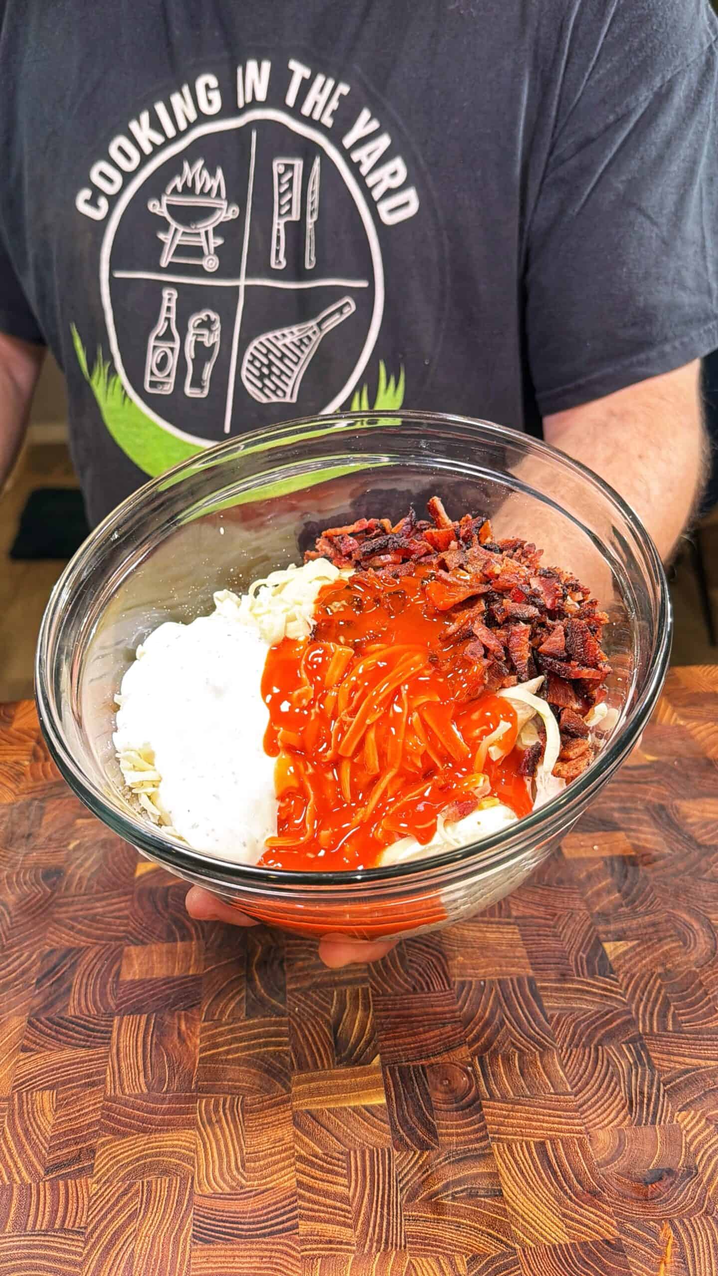 A person in a Cooking in the Yard shirt holds a glass bowl with sour cream, shredded cheese, bacon bits, and hot sauce on top of noodles, above a wooden surface.