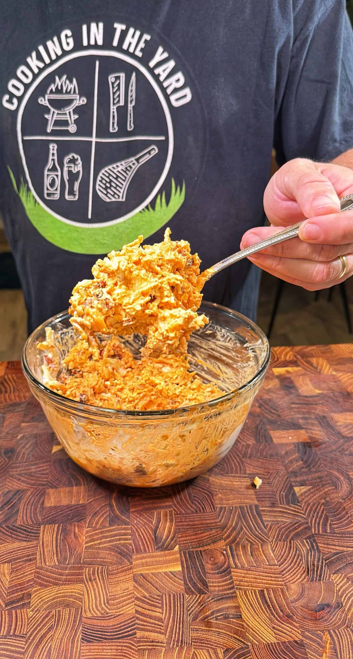 A person wearing a Cooking in the Yard t-shirt holds a forkful of a cheesy, mixed dish from a glass bowl on a wooden table.