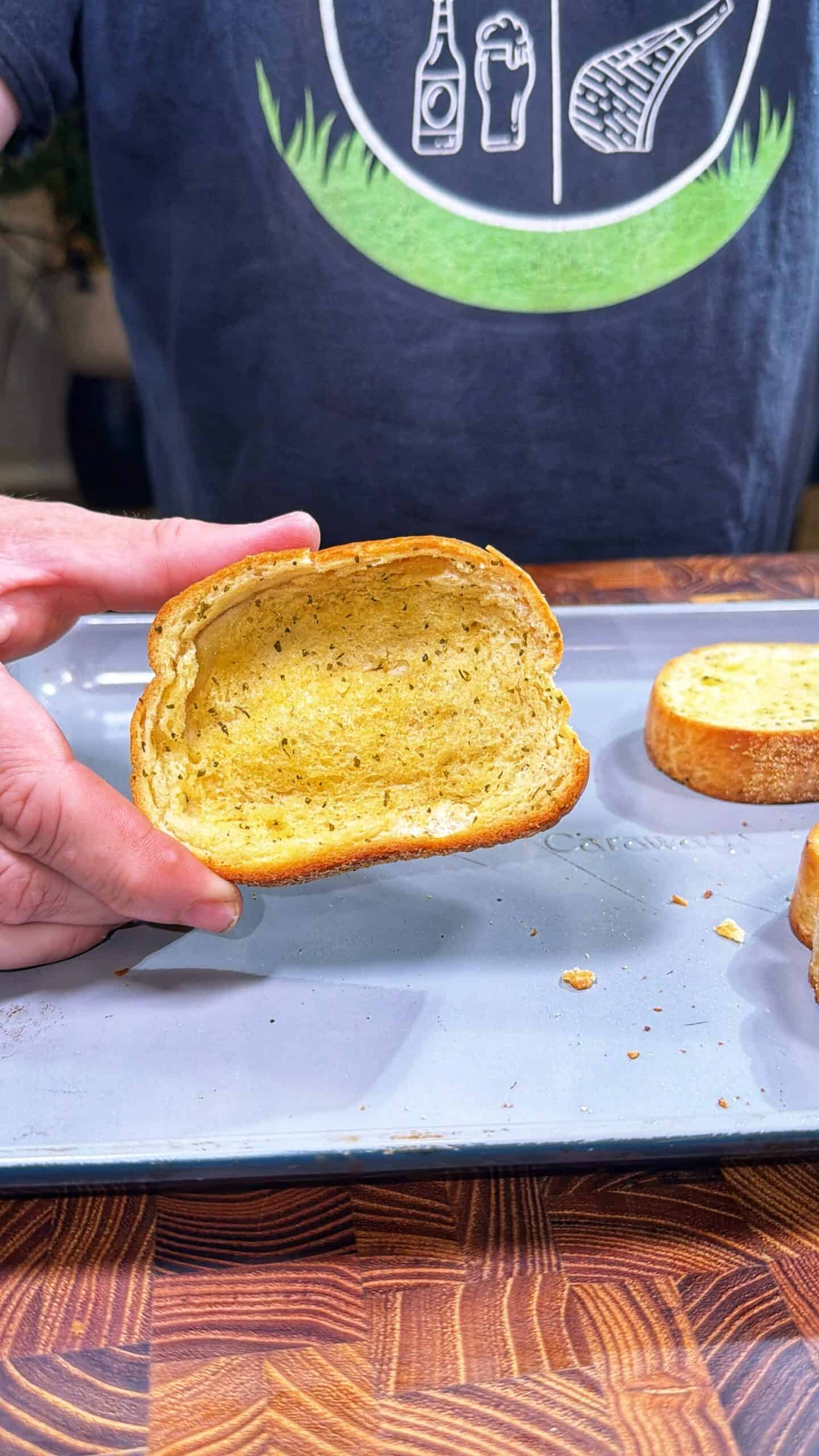 A person holds a slice of seasoned bread above a baking tray with more slices. The person is wearing a dark shirt with white graphic designs near the top.