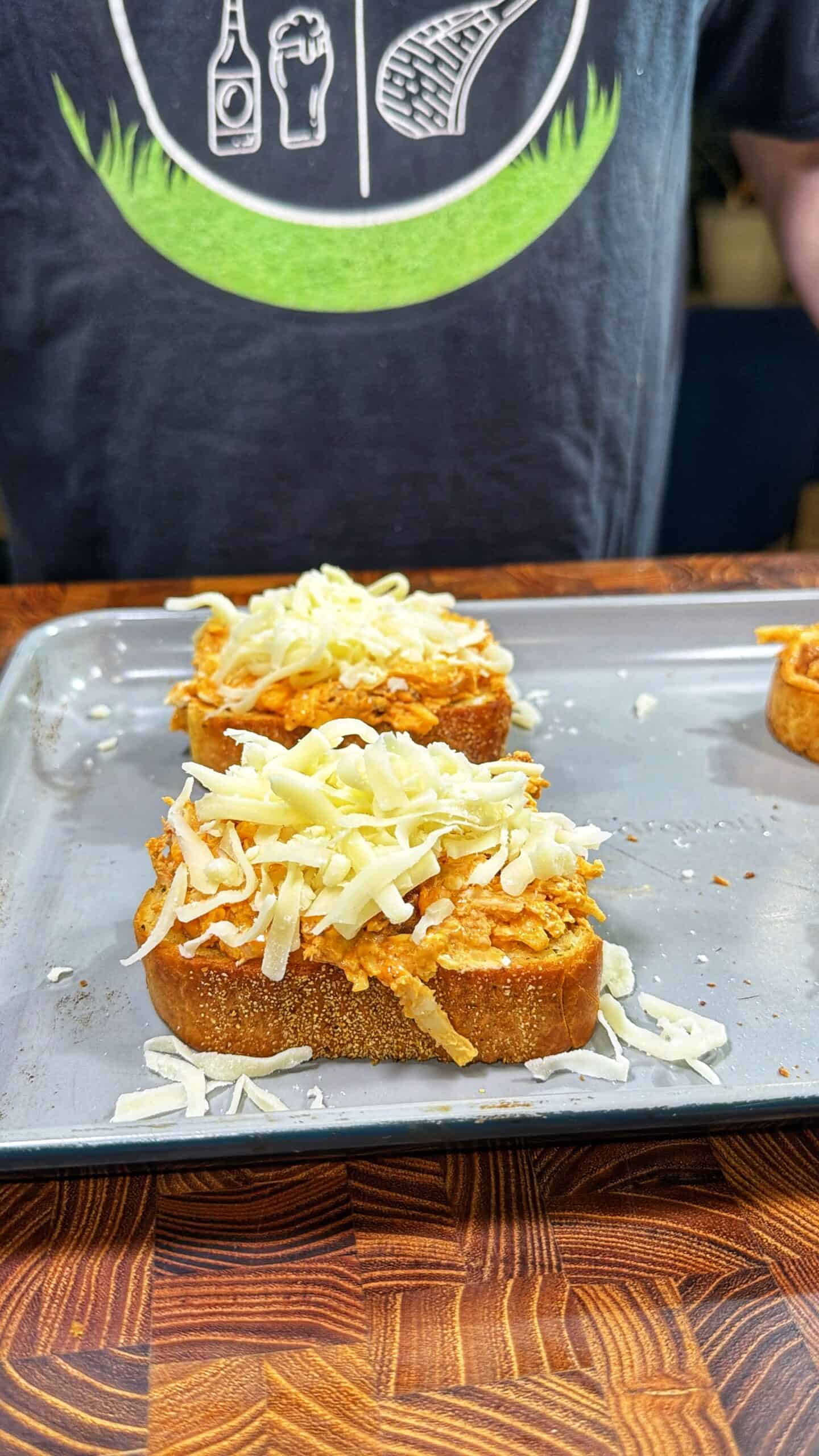Two slices of bread topped with a spread and shredded cheese are placed on a baking sheet. Behind them, a person wearing a dark T-shirt with food-related graphics stands at a wooden countertop.