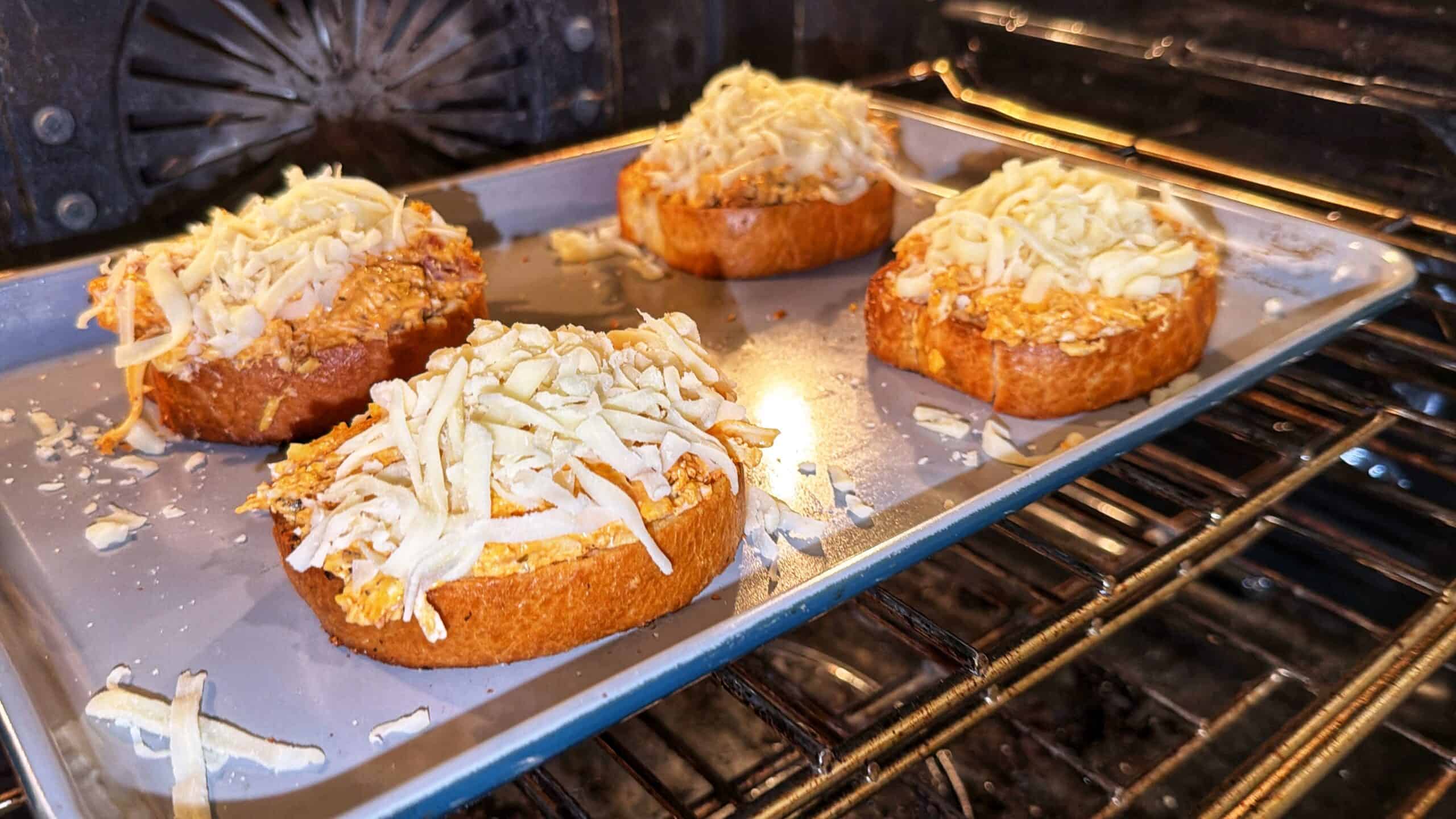 Four slices of bread topped with shredded cheese are on a baking tray inside an oven, beginning to melt and toast.