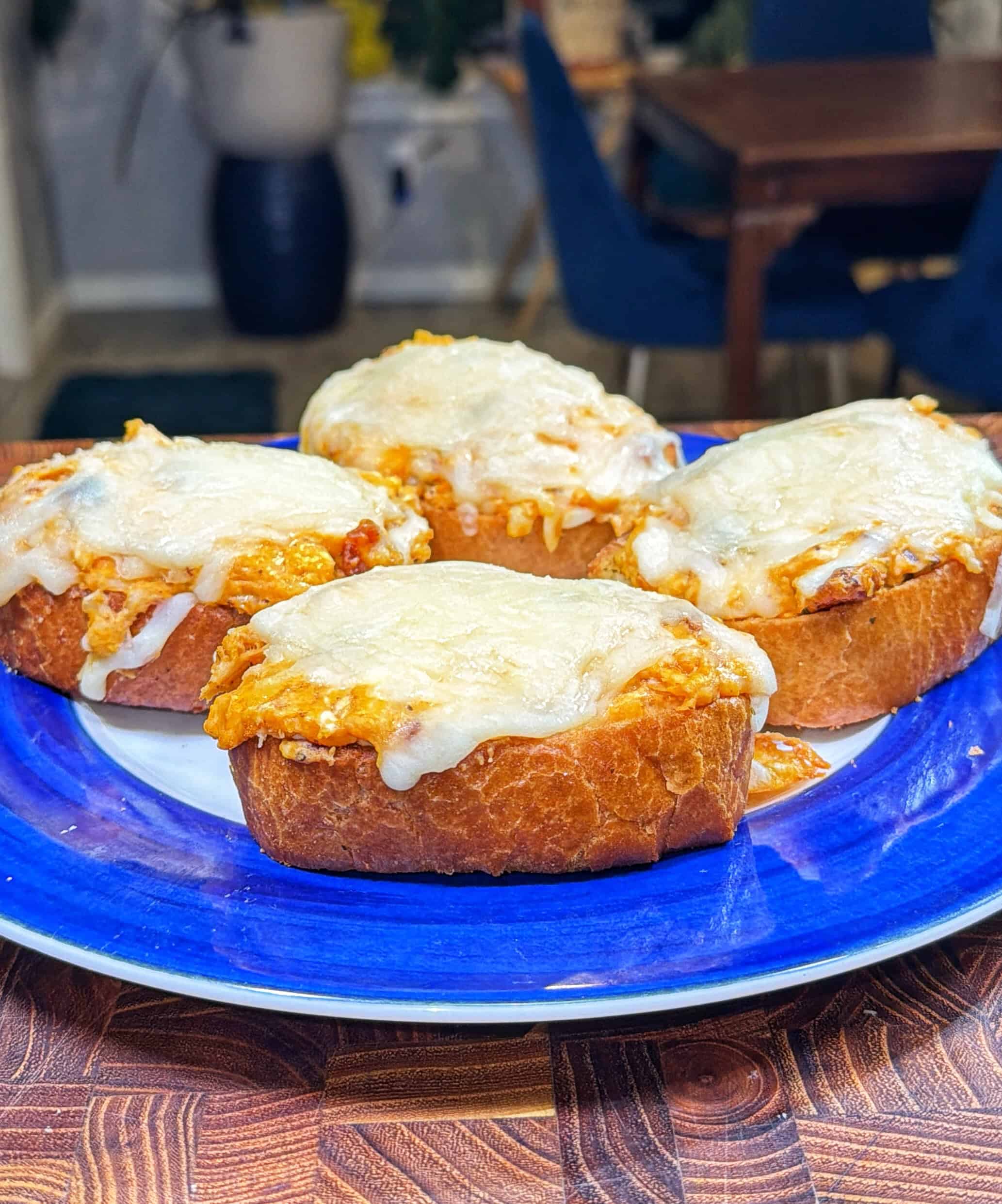 Four slices of toasted bread topped with a cheesy, orange filling and melted white cheese are arranged on a blue and white plate, sitting on a wooden surface. A dining area is visible in the background.