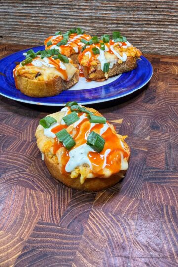 Open-faced sandwiches topped with melted cheese, buffalo sauce, ranch dressing, and chopped green onions, displayed on a wooden surface. Three sandwiches are on a blue plate in the background; one is in the foreground.
