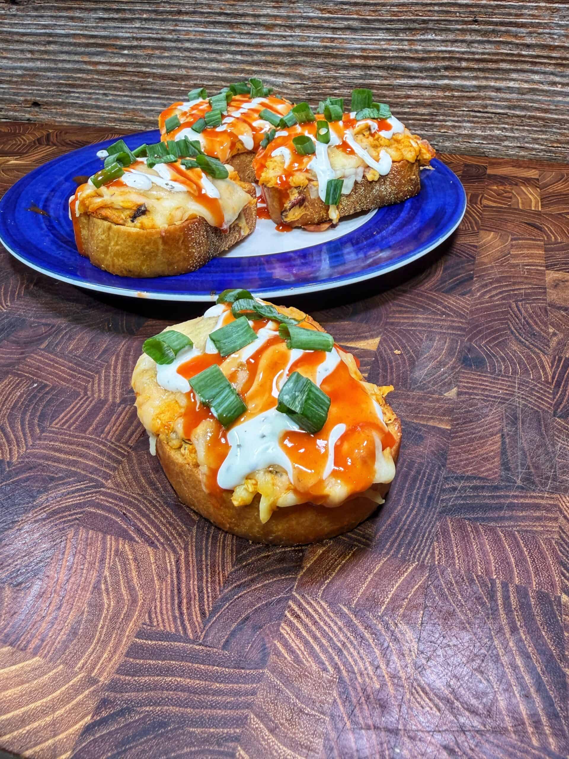 Open-faced sandwiches topped with melted cheese, buffalo sauce, ranch dressing, and chopped green onions, displayed on a wooden surface. Three sandwiches are on a blue plate in the background; one is in the foreground.
