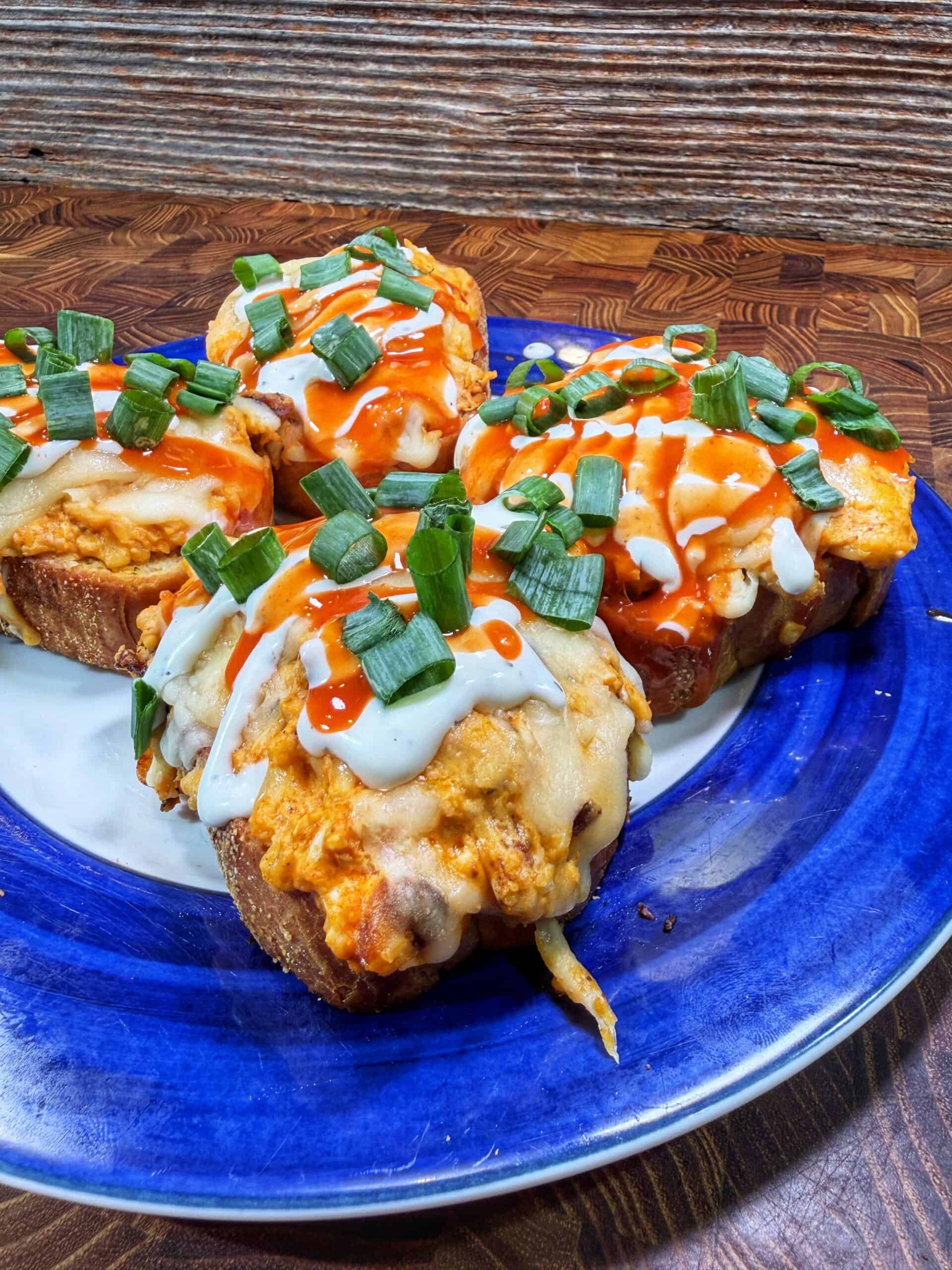 Four pieces of toasted bread topped with melted cheese, buffalo chicken, chopped green onions, and drizzled with ranch and hot sauce, served on a blue and white plate on a wooden surface.