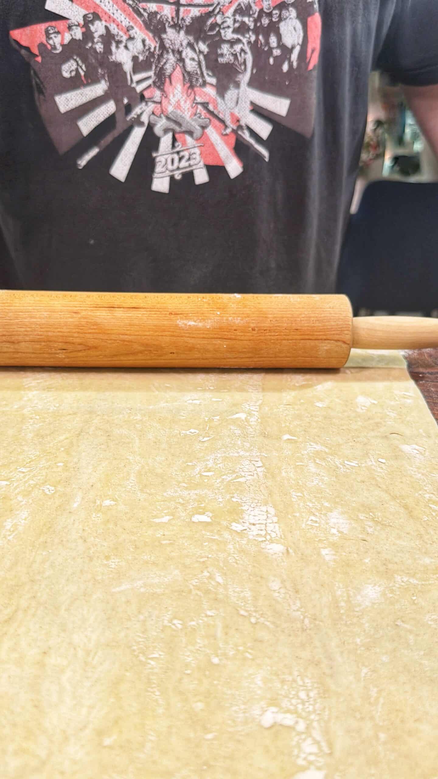 A person wearing a dark graphic T-shirt stands behind a rolling pin and a large sheet of dough on a floured surface, preparing to roll it out.