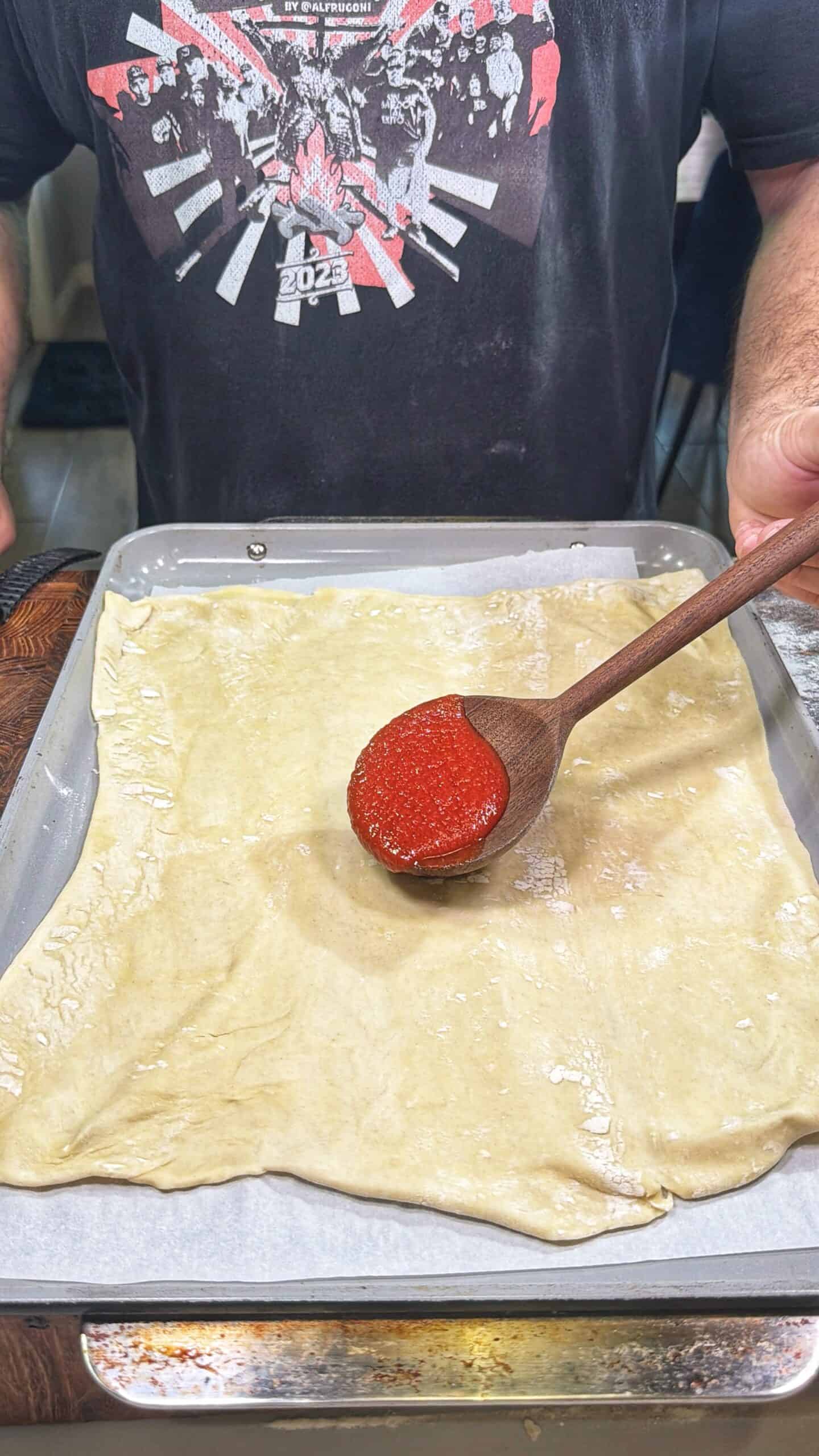 A person spreads tomato sauce with a wooden spoon onto a rectangular sheet of dough on a baking tray lined with parchment paper. The person is wearing a black graphic t-shirt.