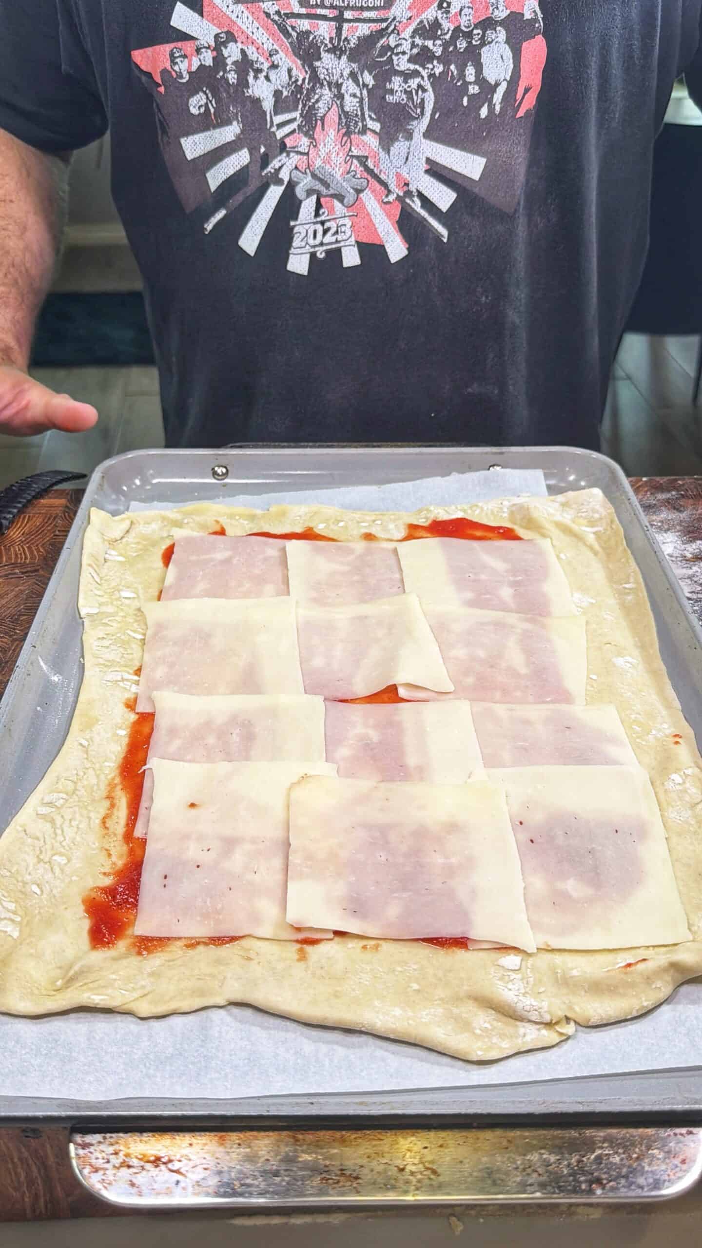 A person wearing a dark graphic t-shirt stands behind a baking tray with rolled-out dough, topped with tomato sauce and overlapping slices of cheese and deli meat.