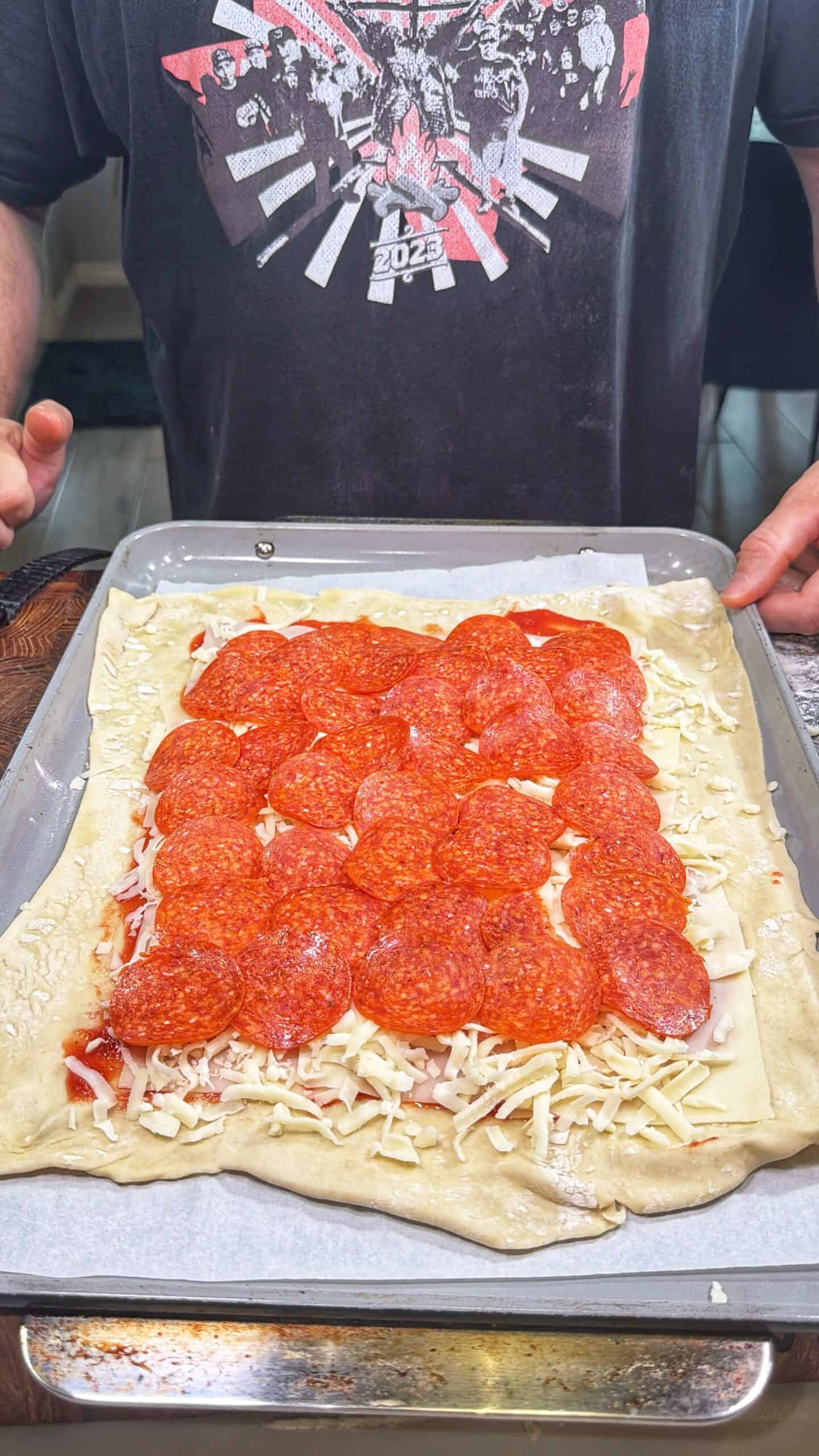 A person stands behind a baking sheet with uncooked rectangular pizza dough topped with tomato sauce, shredded cheese, and neatly arranged pepperoni slices. The person is wearing a dark graphic T-shirt.