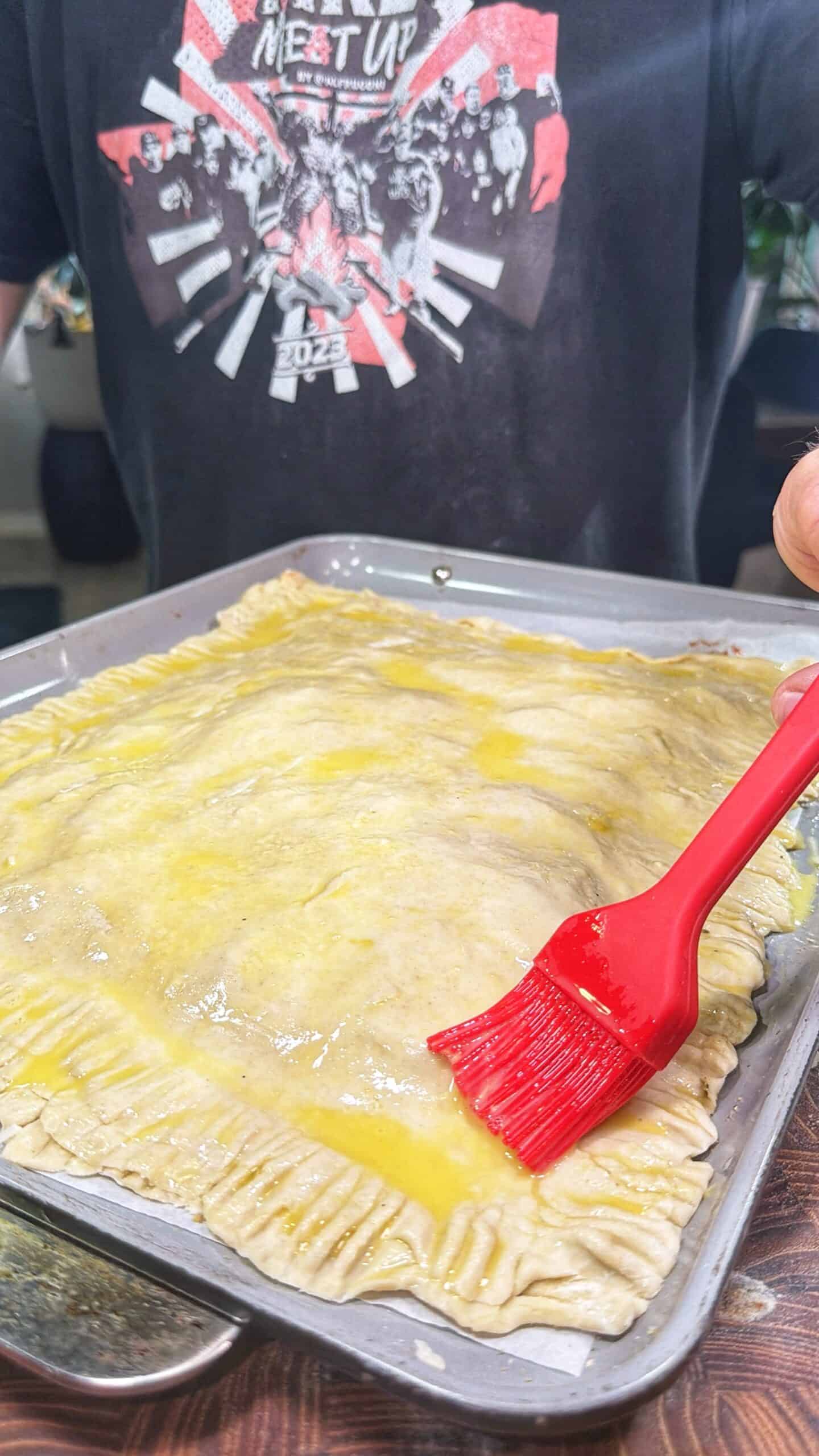 A person uses a red brush to apply egg wash to a large rectangular pastry with crimped edges on a baking tray. The person is wearing a black graphic t-shirt in the background.