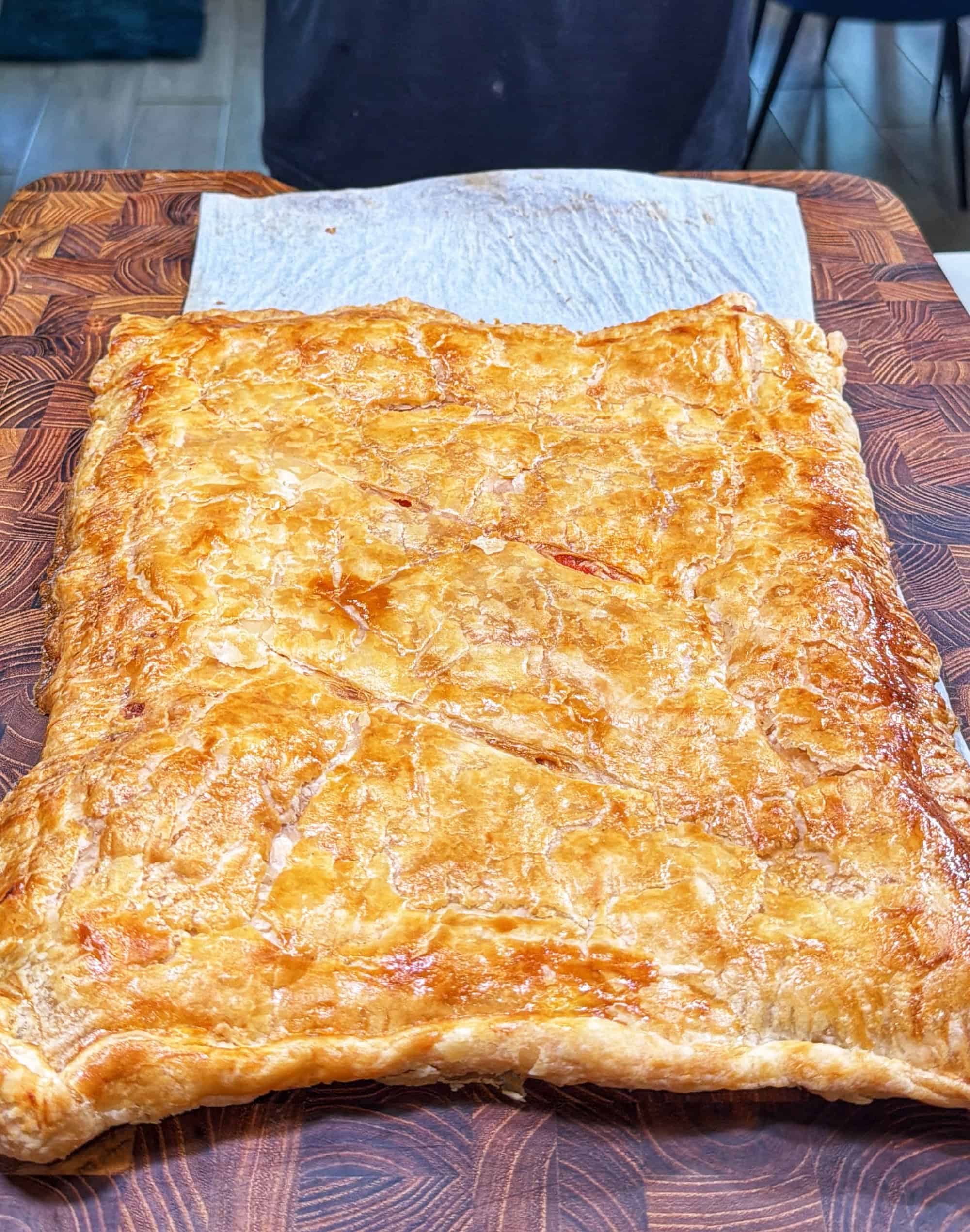 A large, golden-brown rectangular pastry with a flaky crust sits on a patterned wooden cutting board, partially on a piece of parchment paper.