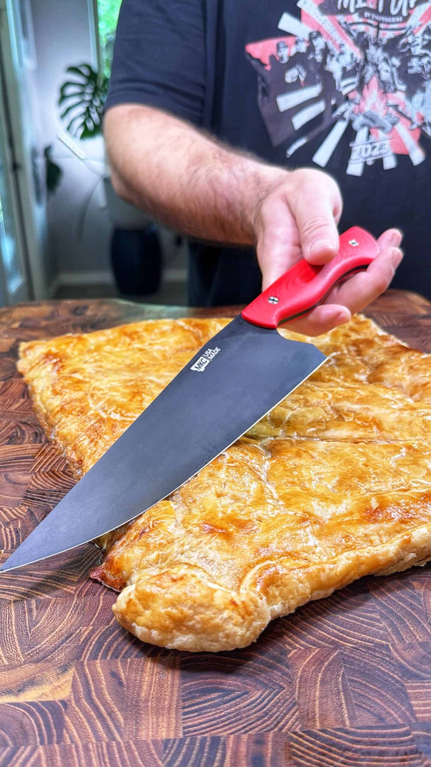 A person holding a large knife with a red handle is about to cut into a golden-brown, flaky pastry resting on a wooden cutting board. The persons torso and arm are visible, and they are wearing a dark t-shirt.
